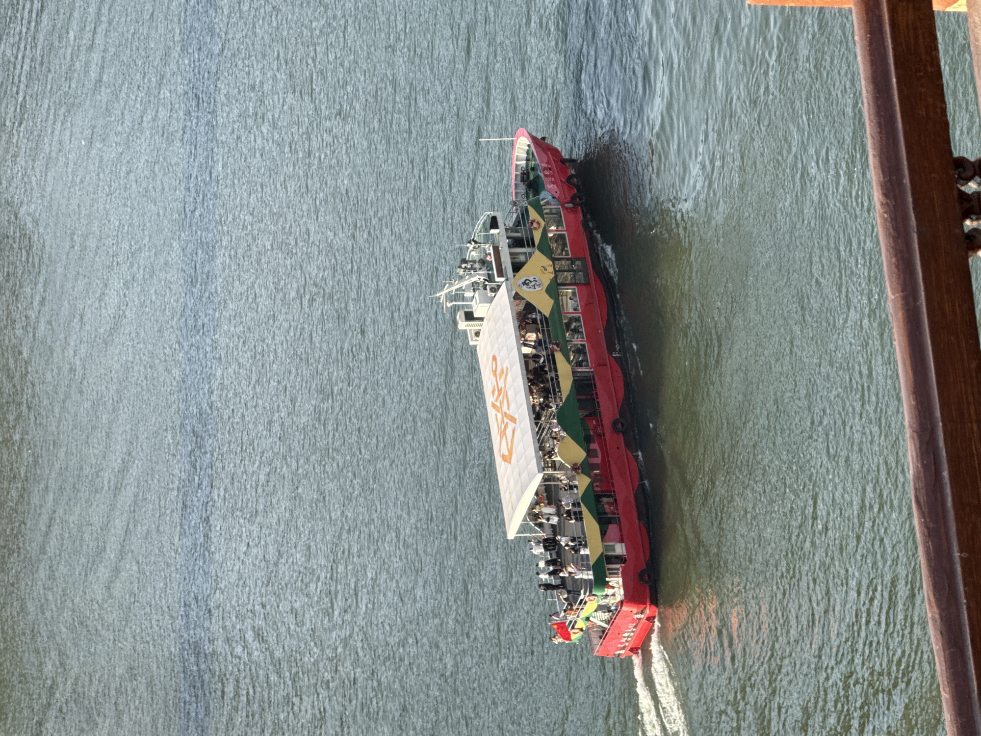 A large, red passenger boat is captured moving across a body of water under bright, clear daylight. The boat features multiple decks, with a prominent white canopy spanning the upper deck. The hull is predominantly red, accented with green and yellow decorative patterns along its sides. Numerous people are visible on the boat's upper levels, appearing as passengers, with some standing and others seated. A white wake trails behind the boat's stern, indicating it is actively moving.
Large Chinese characters are prominently displayed on the white canopy, which read "两江游" (Liǎngjiāng Yóu), translating to "Two Rivers Tour," suggesting the boat operates as a sightseeing vessel.
The water surface exhibits ripples and choppiness, reflecting the bright sunlight. The photograph appears to be taken from a high vantage point, possibly a bridge or elevated structure, looking down onto the river. A brown, wooden railing is partially visible along the right edge of the frame, further suggesting an elevated perspective. The scene depicts a sunny day in Yuzhong District, China.