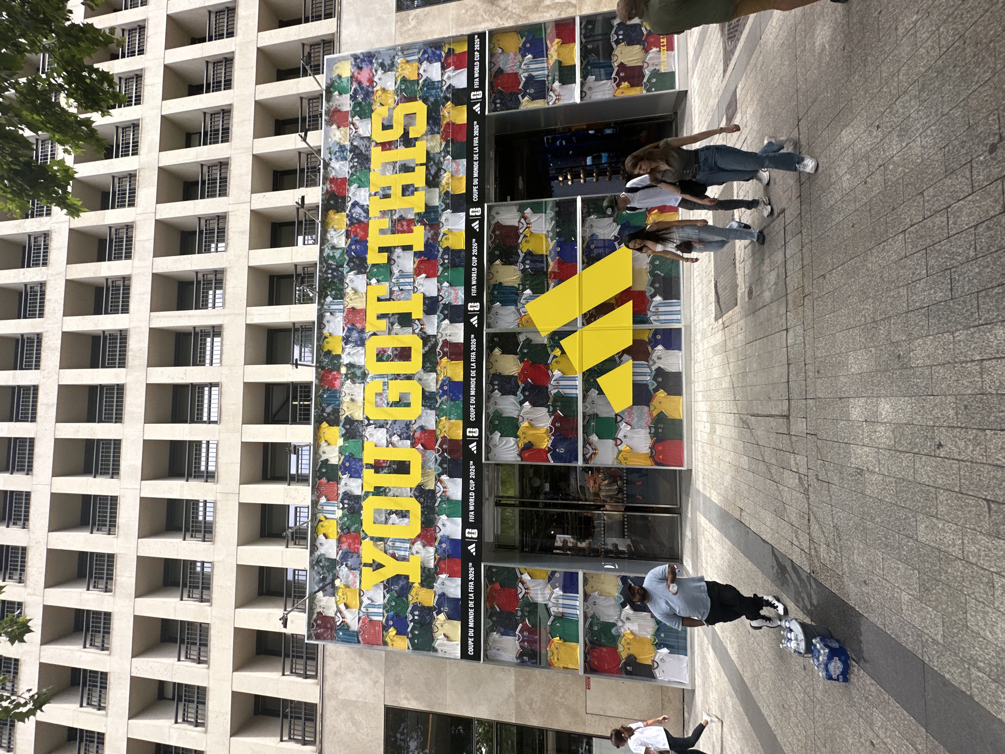 In Paris, France, on a bright day, an Adidas store's facade is prominently displayed, featuring a large graphic with the words "YOU GOT THIS" in yellow letters against a backdrop of numerous football jerseys in various colors. The store itself has large glass doors and windows, and the surrounding building has a repetitive architectural design with many balconies. Several people are walking on the sidewalk in front of the store. One man stands near a bucket containing bottles of water and cases of bottled water. Another group of three young people, two women and one man, are walking past the store. A woman in white is walking on the far left of the image. The overall scene captures a bustling urban environment with a focus on sports merchandising.