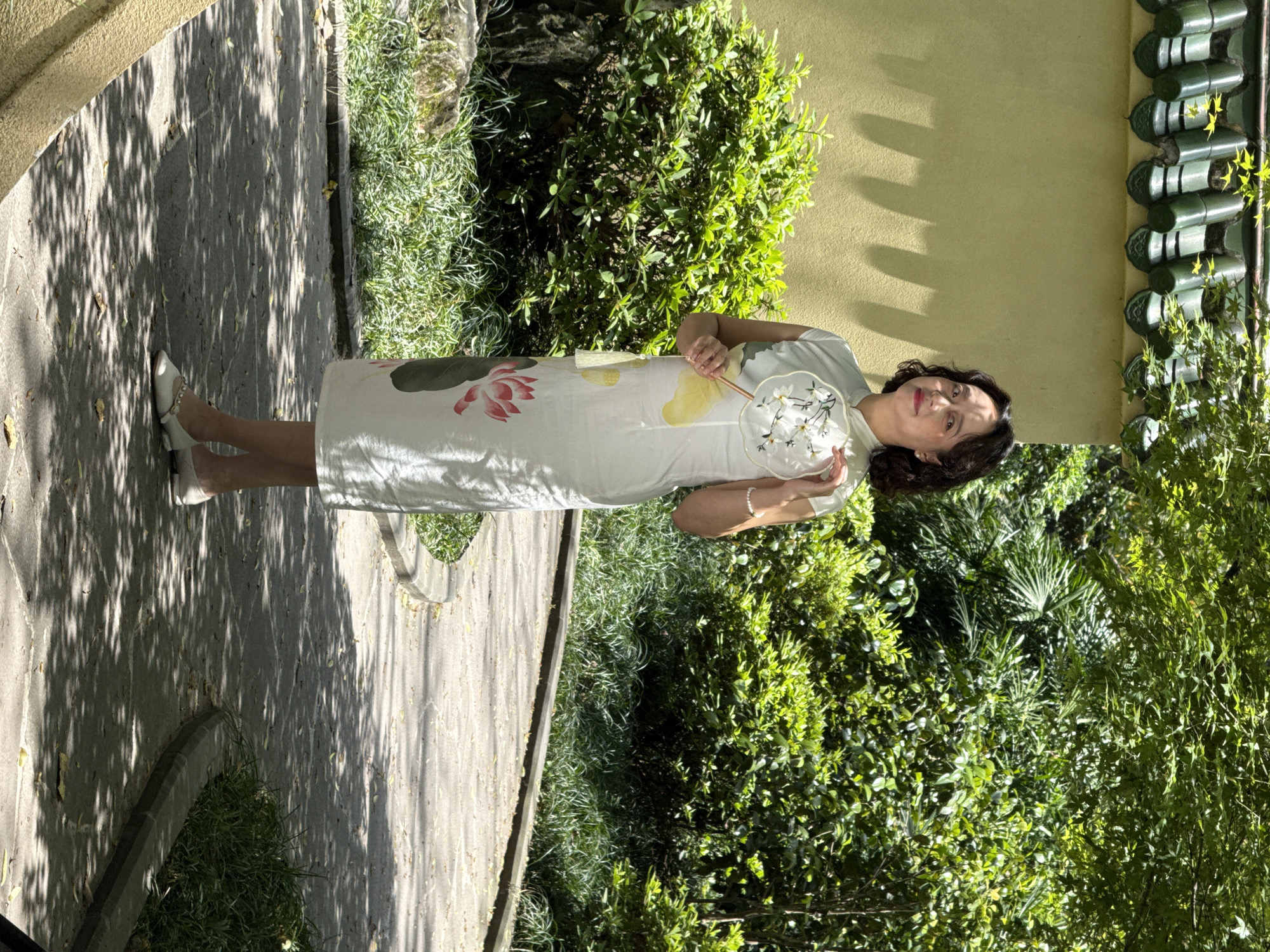 A middle-aged woman stands in a garden in the Yuzhong District of China. She wears a light-colored, traditional Chinese dress with floral motifs and white low heels, and holds a round fan decorated with white flowers. The woman has dark hair and is smiling. She is positioned between a stone path with dappled sunlight and a lush garden with a variety of greenery, including bushes and trees. To the right, a yellow wall is partially visible with traditional green roofing tiles.