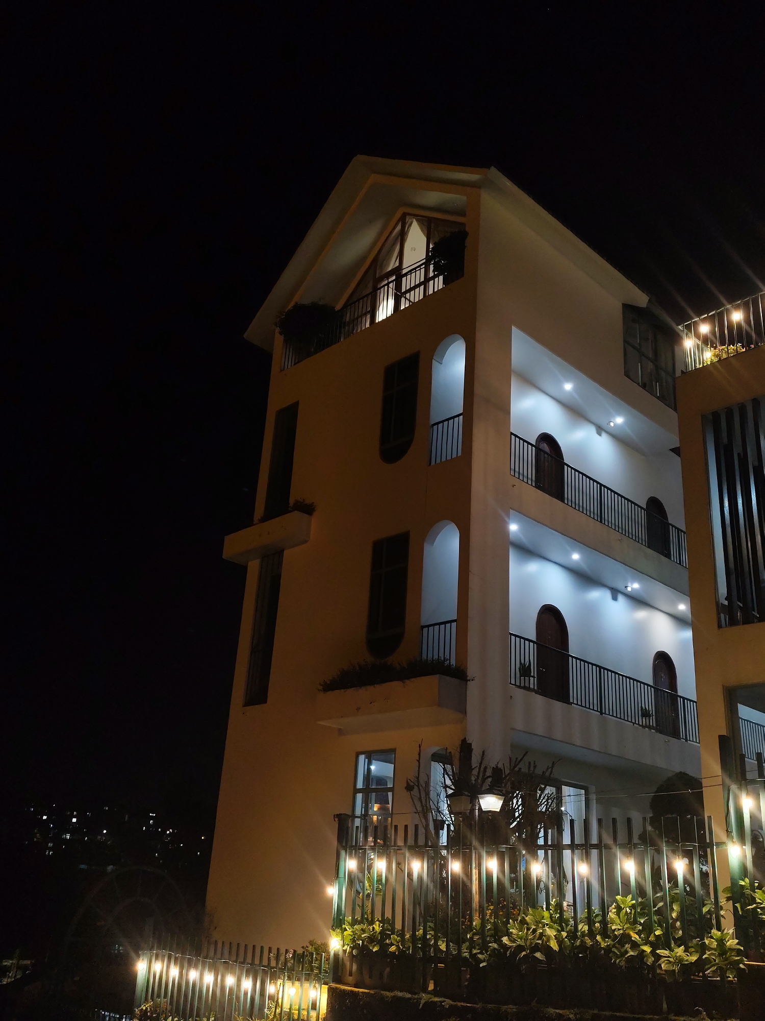 This is a nighttime exterior shot of a modern, multi-story building, likely a hotel or guesthouse, situated on a hillside in Da Lat, Vietnam. The building's architecture features a combination of cream-colored and light yellow walls with multiple balconies and arched doorways. Each balcony is protected by black metal railings. The interior lights are on, illuminating the balconies and doorways with a warm glow. String lights are visible along the fence in the foreground, casting a warm light on the lush green plants below. The background is dark, with the faint glow of distant city lights suggesting its location within Da Lat. The scene is calm and quiet, typical of a serene evening in the mountainous city.