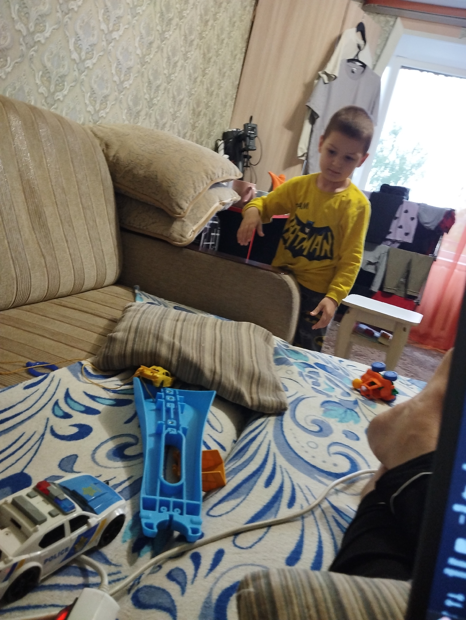 The image shows a boy wearing a yellow Batman shirt standing in a room. He appears to be playing with toys on a bed. A couch is visible in the foreground. The room appears to be a bedroom or living area, with patterned wallpaper and a window in the background. There are clothes hanging on a rack near the window. A small white stool is also visible. The overall impression is that of a domestic setting. No specific details about Kingisepp, Russia, can be inferred from the image.