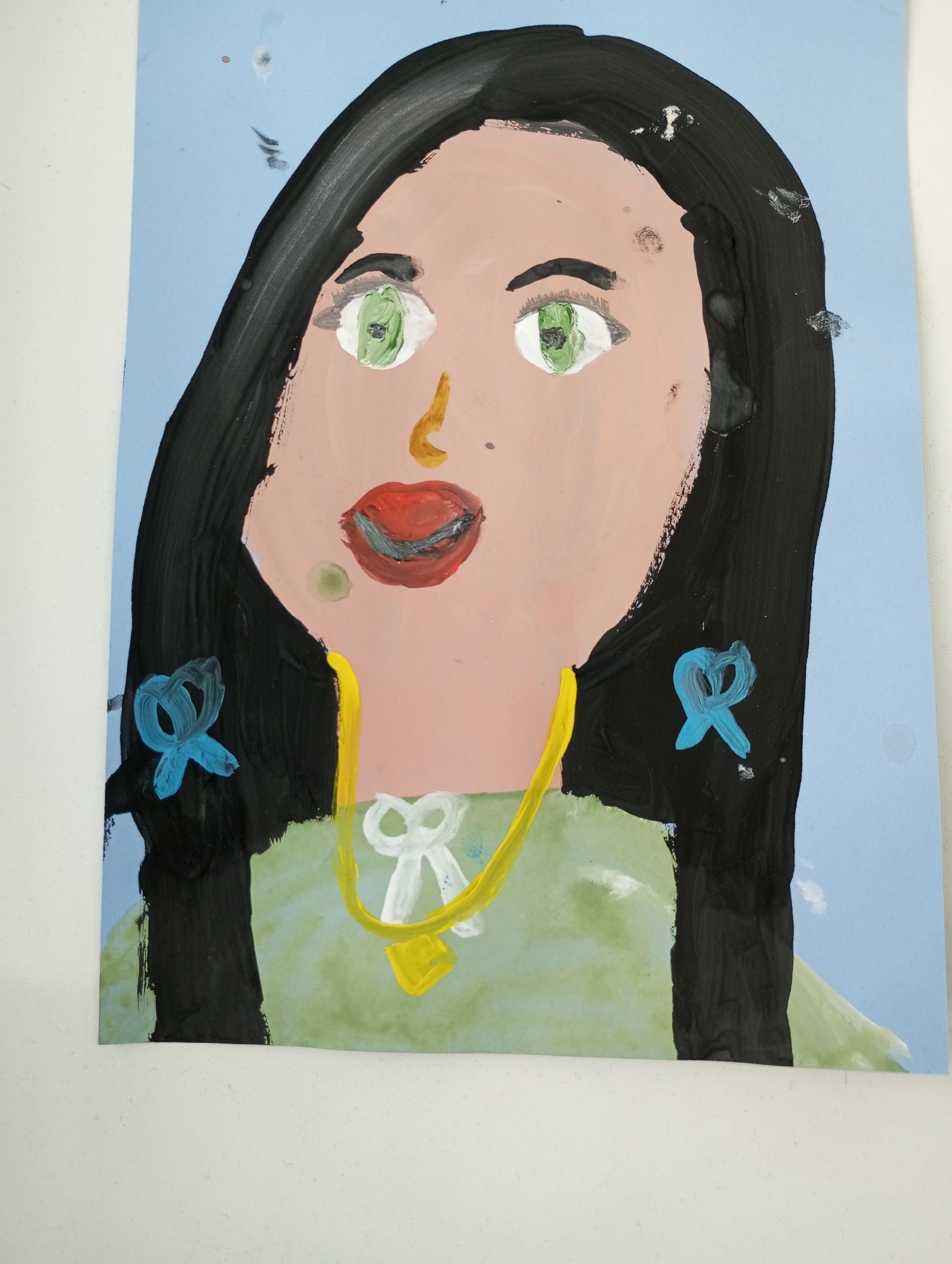 A child's painting on light blue paper depicts a portrait of a woman. The woman has a pinkish-tan face, green eyes, red lips, and black hair with blue ribbons. She is wearing a green shirt and a yellow necklace with a white bow and a yellow pendant. The background is plain light blue. No location in Pervomaysky, Russia is indicated.