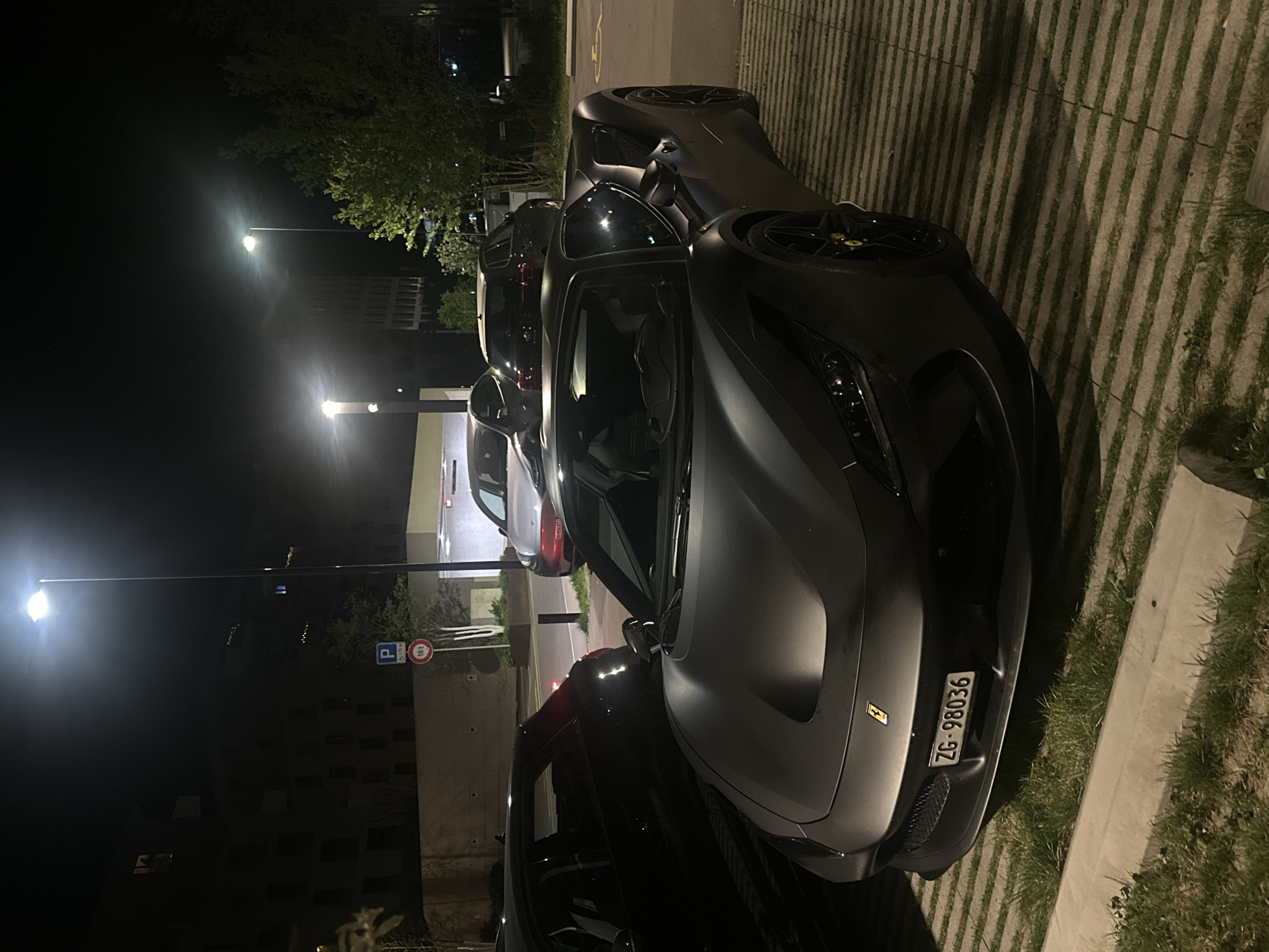 The image captures a nighttime scene in what appears to be a parking area in Dübendorf, Switzerland. The primary subject is a dark, matte-finished sports car, likely a Ferrari, parked on a permeable pavement surface featuring concrete strips interspersed with grass. The car's sleek design and dark wheels with yellow accents (possibly brake calipers or center caps) are visible.
The time of day is clearly night, with multiple streetlights illuminating the scene, casting reflections and shadows. The background shows other parked vehicles and distant buildings, barely visible through the darkness, along with some trees.
A prominent detail is the Swiss license plate on the rear of the main car, which reads "ZG-98036". "ZG" corresponds to the Canton of Zug, providing a specific geographical cue within Switzerland, aligning with the provided location context. In the mid-ground, a blue road sign with a white "P" symbol for parking is visible, next to a red circular sign with "18t" inside, likely indicating an 18-ton weight limit. Another small, red circular sign is partially obscured.
No people are visible, and no active event is taking place; the cars are stationary. The overall atmosphere is quiet, typical of a parked area at night.