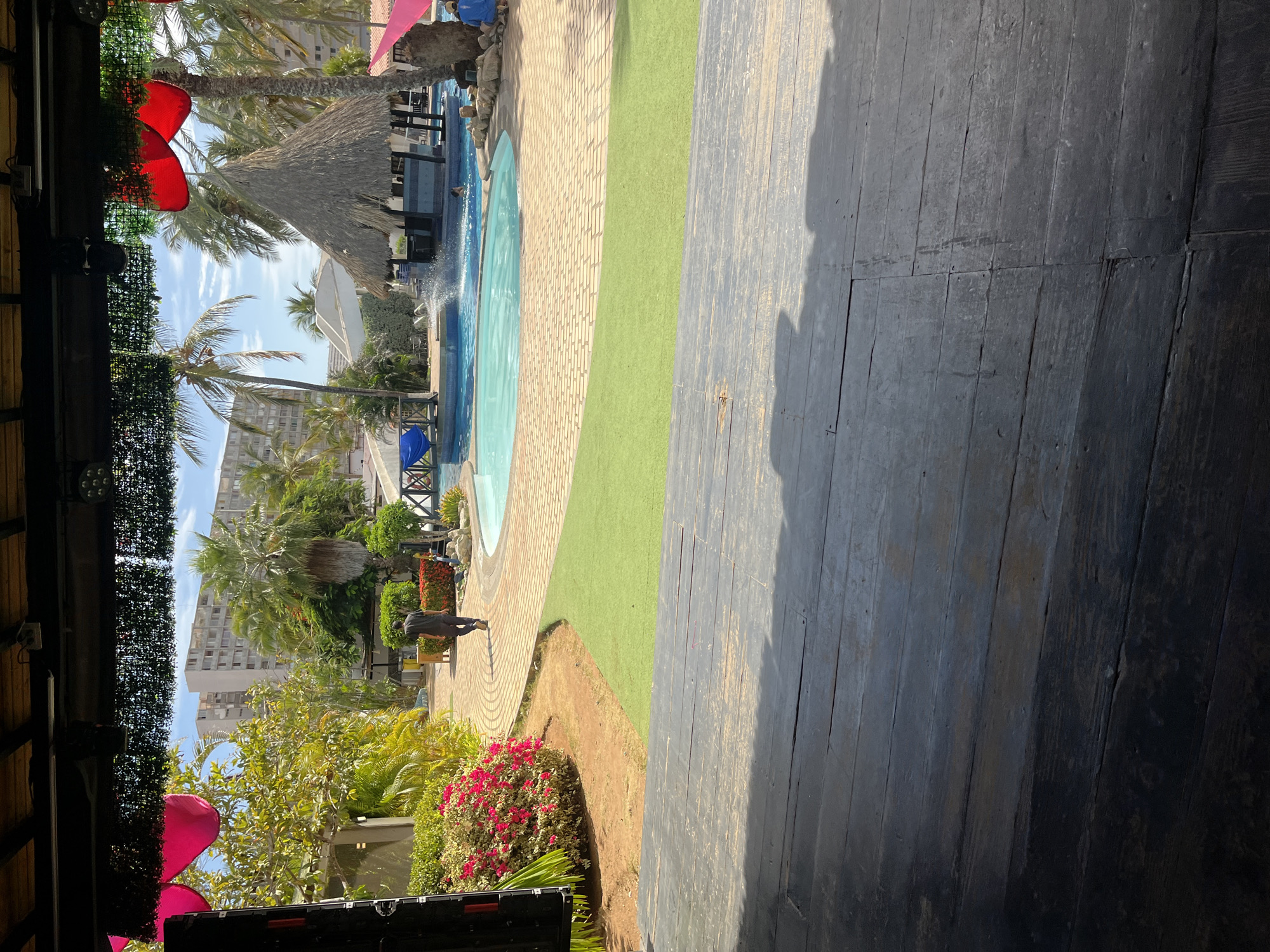 The image shows an outdoor scene in what appears to be a resort or recreation area. There is a pool with a thatched-roof structure (likely a palapa) nearby. Palm trees and various plants, including some with red flowers, are visible. A multi-story building can be seen in the background, indicating an urban setting. A person in dark clothing is walking by the poolside. The scene is partially viewed from under a structure, possibly a covered patio or deck, with wooden planks in the foreground and some stage lighting and decorations visible. The location is suggested to be Porlamar, Venezuela.