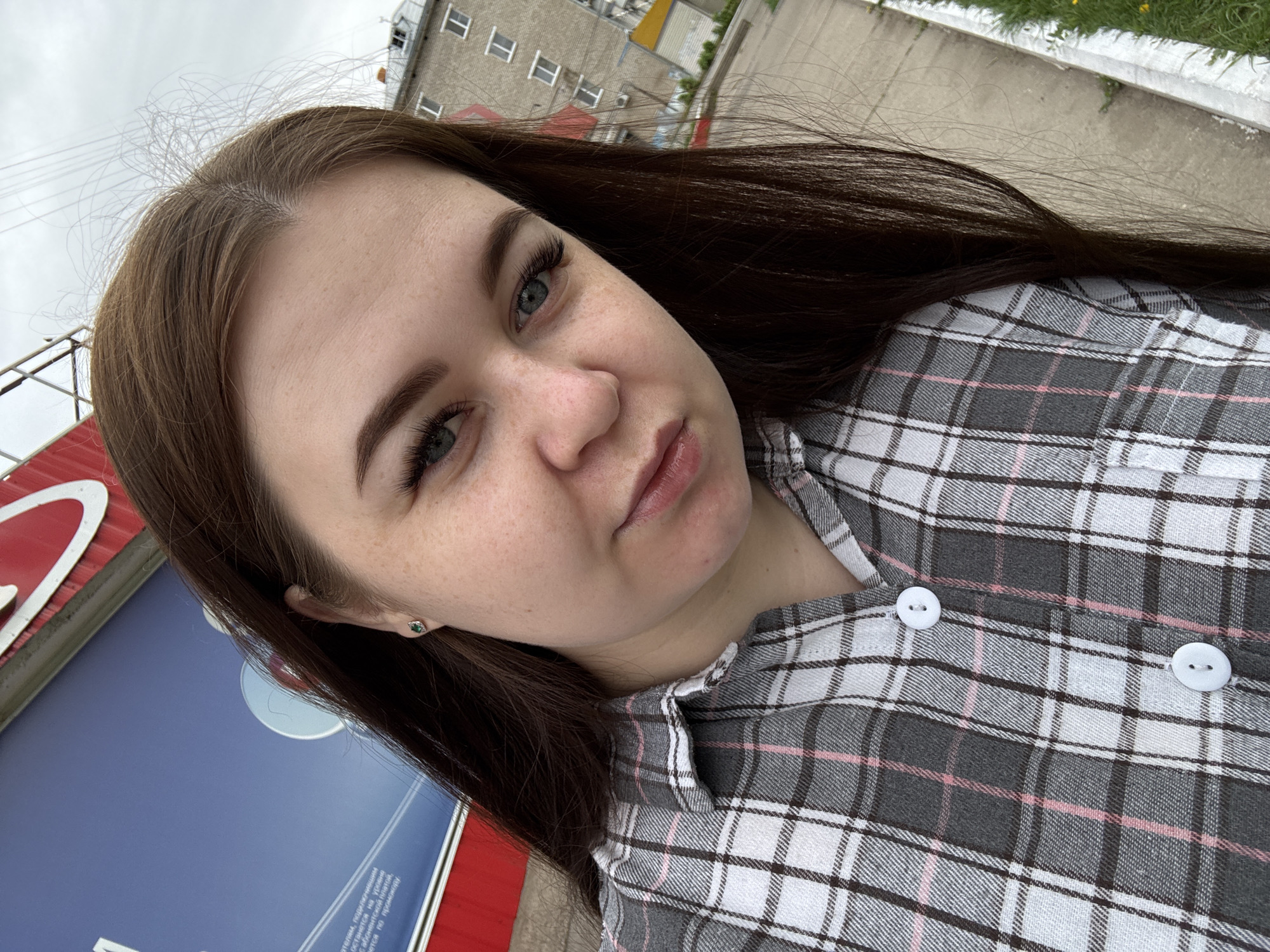 A medium-shot, angled selfie photograph taken outdoors captures a young woman with fair skin and freckles. Her hair is long, straight, and dark brown, falling to her right. She has light eyes, prominent eyebrows, and is wearing eyelash extensions. She is looking directly at the camera with a slight pout. She is wearing a gray, white, and pink plaid button-down shirt. A small, green earring is visible in her left ear.
In the background, there is a gray building with multiple windows, suggesting it is likely an apartment building or office block. The sky appears overcast. Part of a structure with red and blue components is visible to the left, suggesting a sign or advertising panel. A concrete sidewalk is seen at the top right with some grass and vegetation.
This image was taken outside in Chaikovsky, Russia.