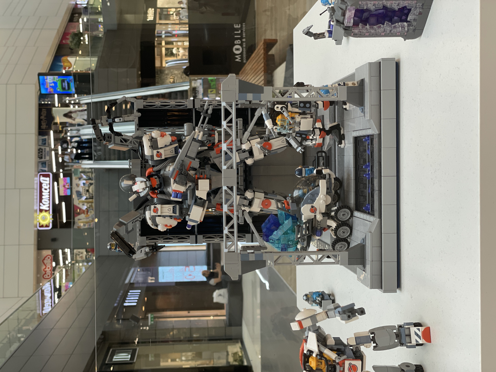 The image shows a display of Lego creations, possibly in a shopping mall. The central subject is a large, grey and white robot figure constructed from Lego bricks. This robot is positioned on a platform with scaffolding, suggesting it is being assembled or repaired. Several smaller Lego vehicles and figures are also part of the display, including a white wheeled vehicle with blue translucent blocks, and a smaller robot or mecha on the left. In the background, a blurred view of a shopping mall interior is visible, featuring escalators, shops with signage like "Komcell" and "Mobile accessories & services," and people walking. The lighting suggests an indoor environment. The display is set up on a white surface, likely a table or counter, with a glass barrier in front. The overall scene depicts a hobbyist display of elaborate Lego models within a commercial setting.