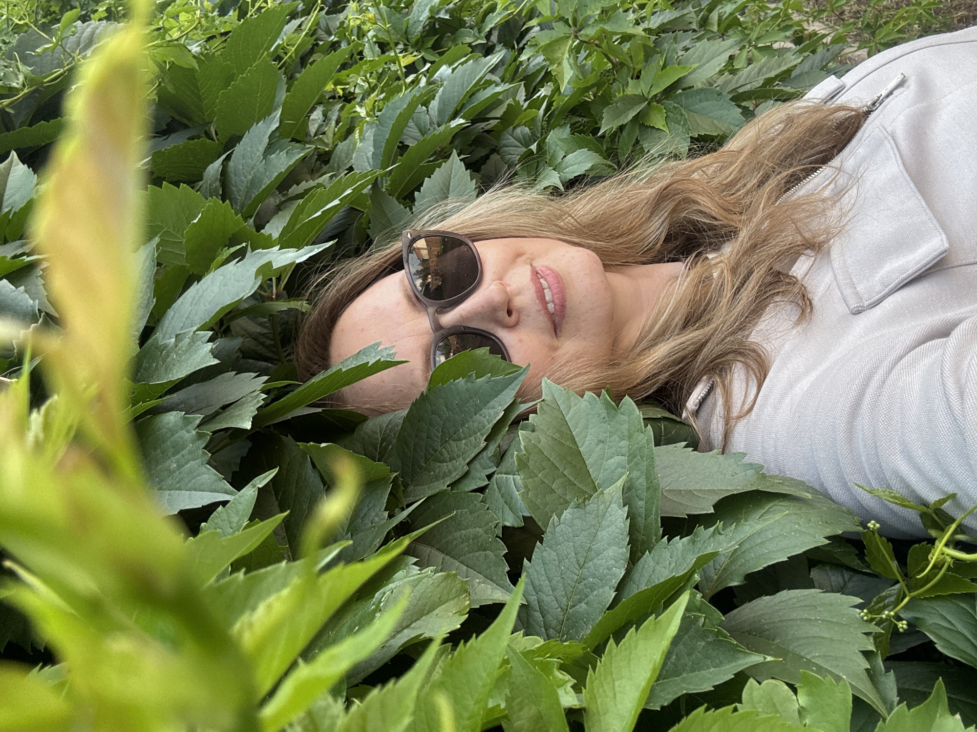A woman with blonde hair and sunglasses is partially obscured by green leaves. She is wearing a light-colored jacket with a zipper. The leaves are dense and appear to be on a wall or fence. The background shows more foliage and a hint of a pathway reflecting in her sunglasses. The location is the city of Chaikovsky, Russia.
