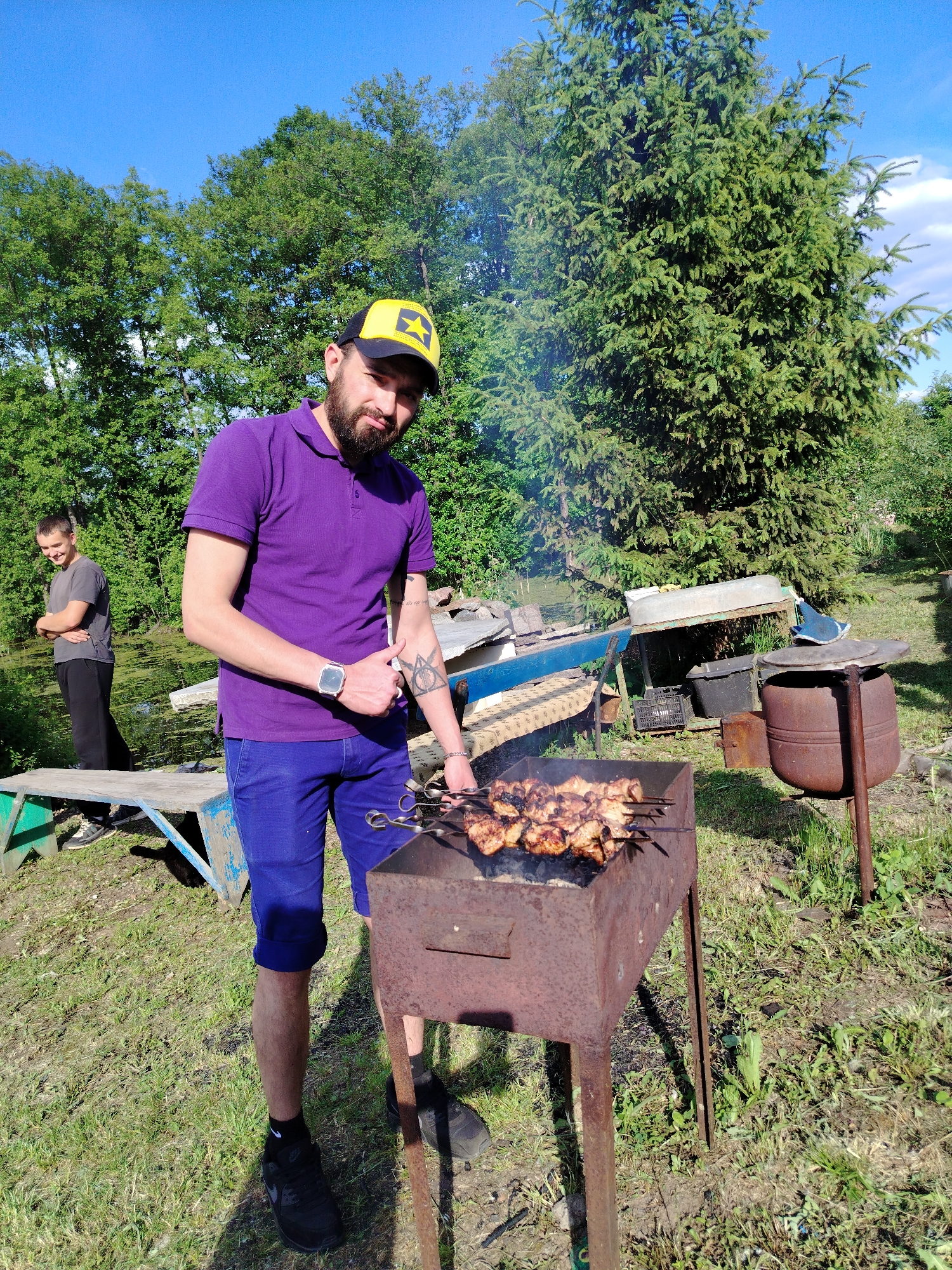 In the city of Noviy, Russia, an outdoor scene unfolds under a clear blue sky with sparse white clouds. In the foreground, a male with a dark beard, a yellow and black baseball cap featuring a yellow star, a purple short-sleeved polo shirt, blue shorts, black socks, and black Nike sneakers, stands next to a rusty, rectangular metal barbecue grill. He holds multiple metal skewers laden with cooked meat over the coals, with smoke rising from the grill. His left forearm displays a geometric tattoo and a silver smartwatch, while his right hand gives a thumbs-up gesture as he looks directly at the camera with a slight smile.
In the mid-ground to the left, a second male with short dark hair, wearing a grey short-sleeved t-shirt and dark pants, stands with his arms crossed, appearing to look downwards or to the side. He is positioned near a wooden bench next to a body of water, which is partially covered by green aquatic vegetation.
The background features dense green foliage, including numerous large deciduous trees on the left and a prominent, tall evergreen tree on the right. Miscellaneous objects are visible between the two individuals and to the right of the main subject: a blue and wooden bench, an overturned light-colored boat or trough resting on a metal frame, black plastic crates, and a large, rusty, pot-like structure on a stand. The ground consists of uneven grass and dirt.