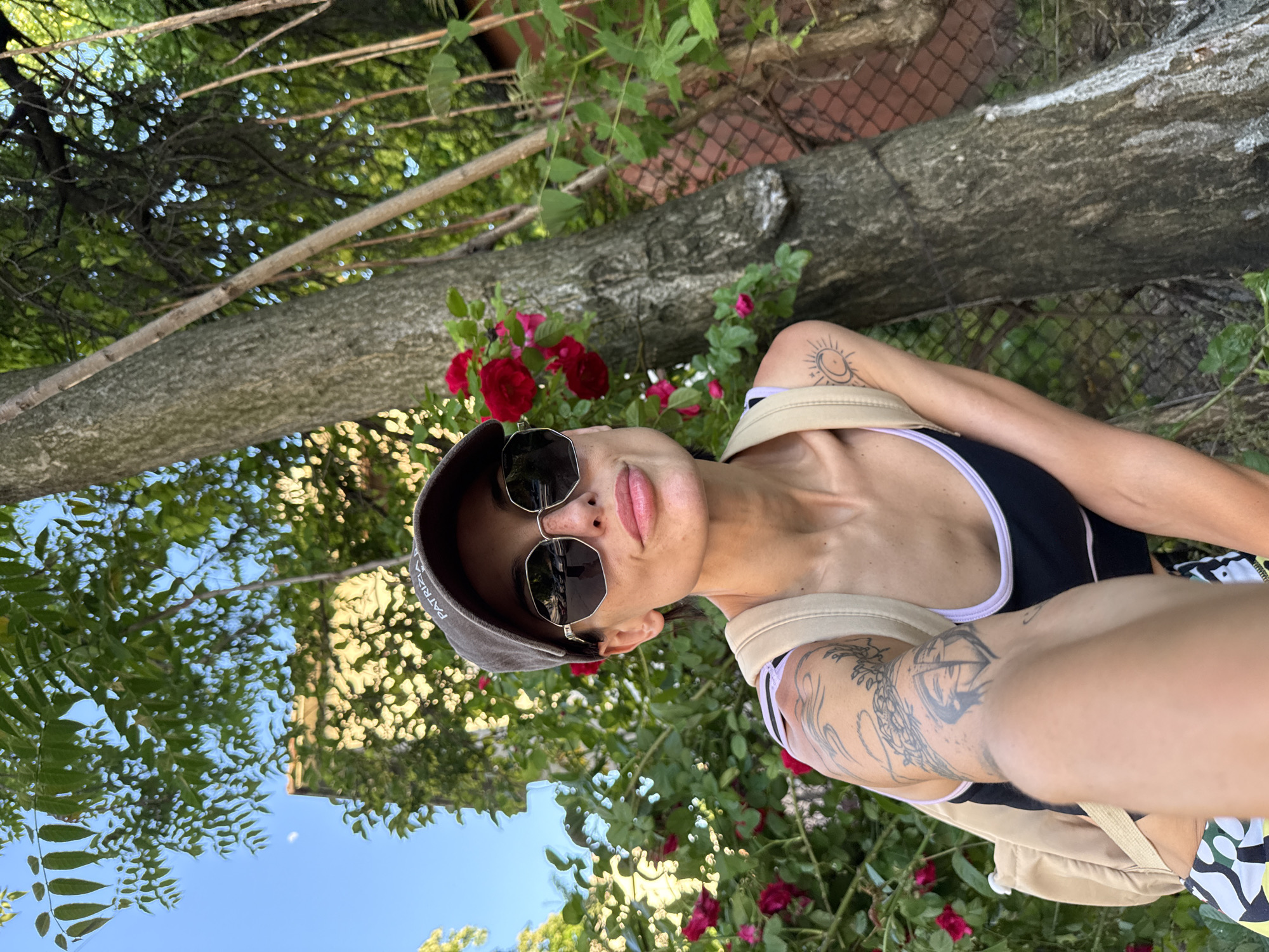 A woman in a black and white tank top, sunglasses, and a cap is taking a selfie in front of a tree with red roses. She has tattoos on her arms and is wearing a backpack. The sky is blue with some green leaves visible. A branch with more roses is to the right of her head, and a chain-link fence behind the branch. It is difficult to determine the specific location in Pleven, Bulgaria from the image alone.