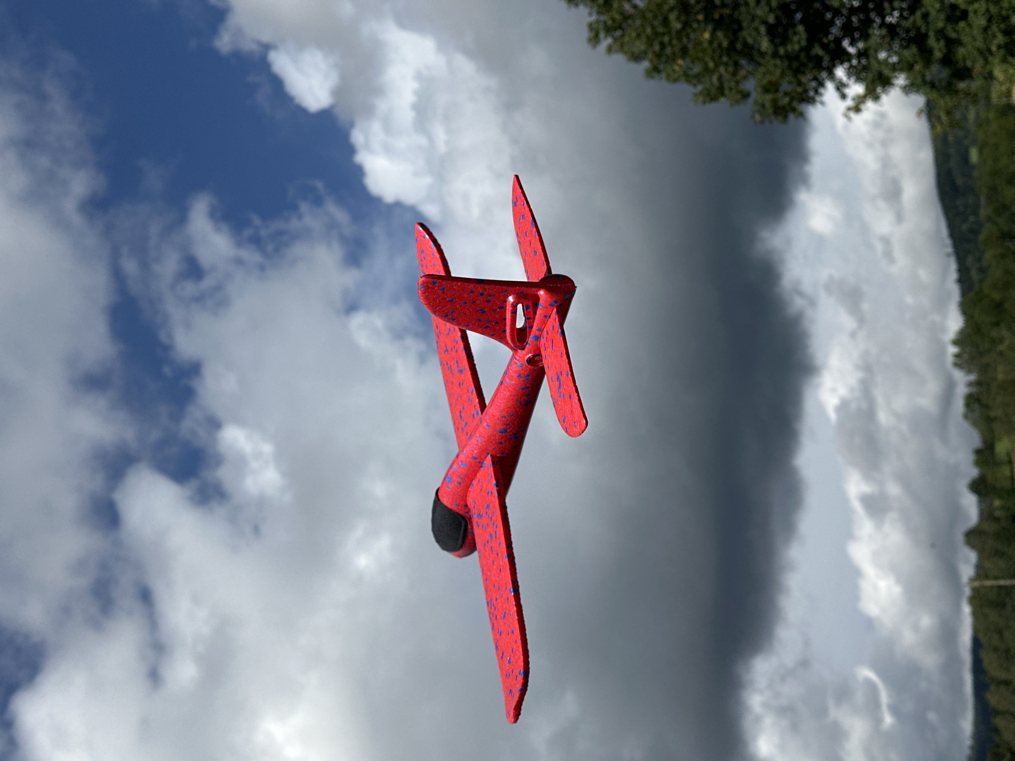 A bright red toy airplane with blue speckles is suspended in mid-air against a backdrop of a dramatic sky. The sky is a mixture of dark, stormy clouds and patches of bright blue sky with fluffy white clouds. The airplane is viewed from a side angle, with its wings extended. In the background, a line of trees and hills is visible under the overcast sky. The image appears to be taken outdoors, likely in a rural or suburban setting. The toy airplane suggests a playful or recreational activity. The location is the city of Wisła, Poland.
