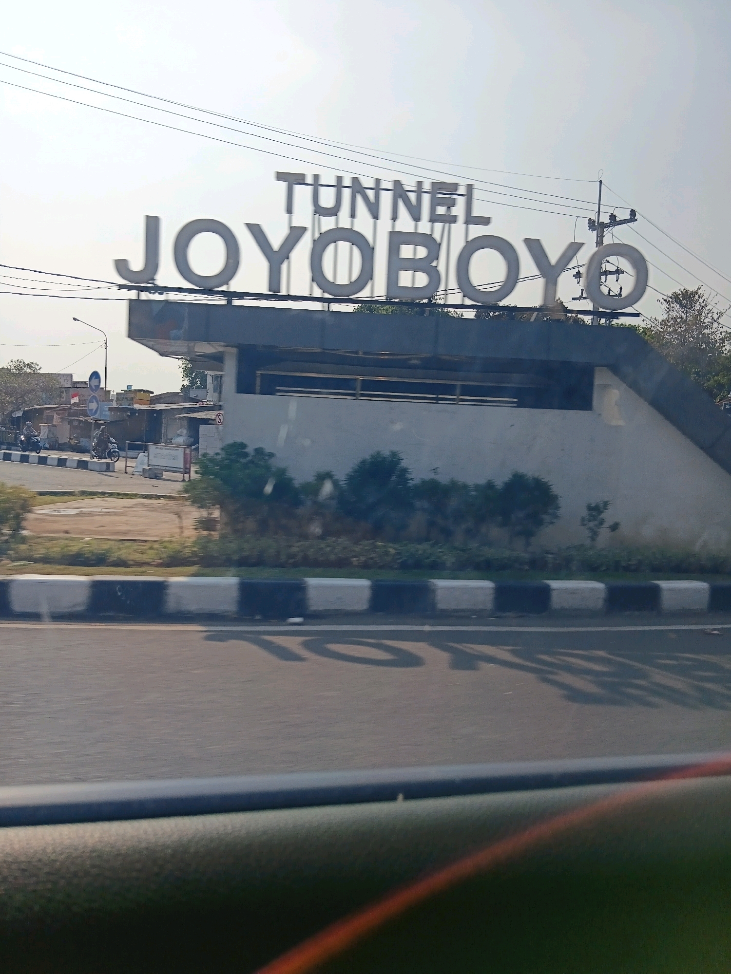 The image captures a view from a vehicle looking towards a tunnel entrance on a bright, possibly overcast day. A large sign above the tunnel reads "TUNNEL JOY OBOYO" in white, three-dimensional lettering. The tunnel entrance is constructed from concrete with a sloping side and a darker, flat roof structure. Below the tunnel, a street is visible with a black and white striped curb. Several small trees and bushes are planted along the side of the road. On the left side of the image, across the street, there are several small buildings with corrugated roofs, a street lamp, and two people on motorcycles. The sky is a pale blue, and power lines are visible crisscrossing overhead. The road in the foreground has shadows from the tunnel sign cast upon it. This scene is located in Surabaya, Indonesia, specifically at the Joyoboyo Tunnel, a significant infrastructure in the city.