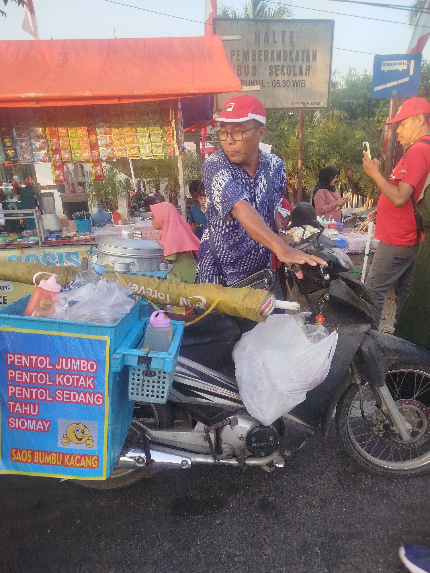 The image depicts an outdoor scene in the city of Burneh, Indonesia, likely near a bus stop, given the presence of a sign that reads "HALTE PEMBERANGKATAN BUS SEKOLAH" (School Bus Departure Stop). The time of day appears to be late afternoon or early evening, as indicated by the warm lighting and shadows.
In the foreground, a man wearing a blue patterned shirt and a red cap is seated on a motorcycle. He is looking to his left, and his right hand is on the motorcycle's handlebars. The motorcycle itself is a dark color, and a white plastic bag is hanging from it. Attached to the side of the motorcycle is a blue cart filled with various food items. Text on the cart indicates it sells "PENTOL JUMBO," "PENTOL KOTAK," "PENTOL SEDANG," "TAHU SIOMAY," and "SAOS BUMBU KACANG." A cartoon emoji with a smiling face and thumbs-up is also visible on the cart.
To the right of the man, another man in a red shirt and red cap is standing and holding a smartphone, appearing to take a photo or video. In the background, several other individuals are visible. A woman in a pink hijab and a green outfit is standing near the motorcycle, looking towards the left. Further in the background, other people are milling around a food stall. The stall is covered by an orange tarp and displays various packaged goods, likely snacks or instant food items. Palm trees and other foliage are visible behind the stall and the bus stop sign, suggesting a tropical environment. The ground is paved with asphalt.
