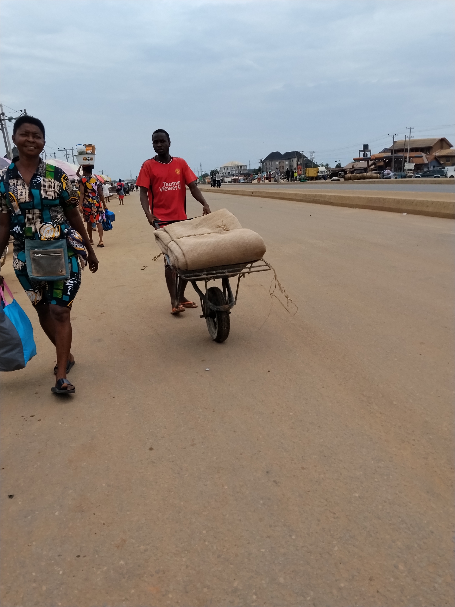The image depicts a street scene in Ura Nta Umuarandu, Nigeria. In the foreground, a woman walks with shopping bags. A man pushes a wheelbarrow loaded with burlap sacks along the side of a road. Other pedestrians are present, with some carrying goods on their heads. Buildings and parked vehicles are visible across the road. Overhead power lines are also seen. The sky is overcast.