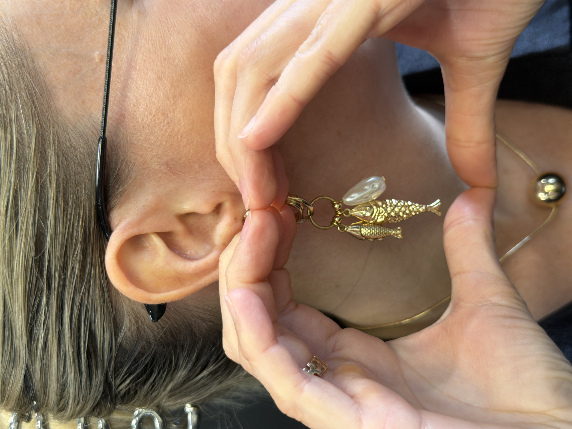 A person is putting a gold fish-shaped earring in their left ear. The earring also features a pearl. The person is also wearing a gold necklace with a large gold ball pendant. The person has blonde hair, and the photo is taken close-up. The location is unknown.