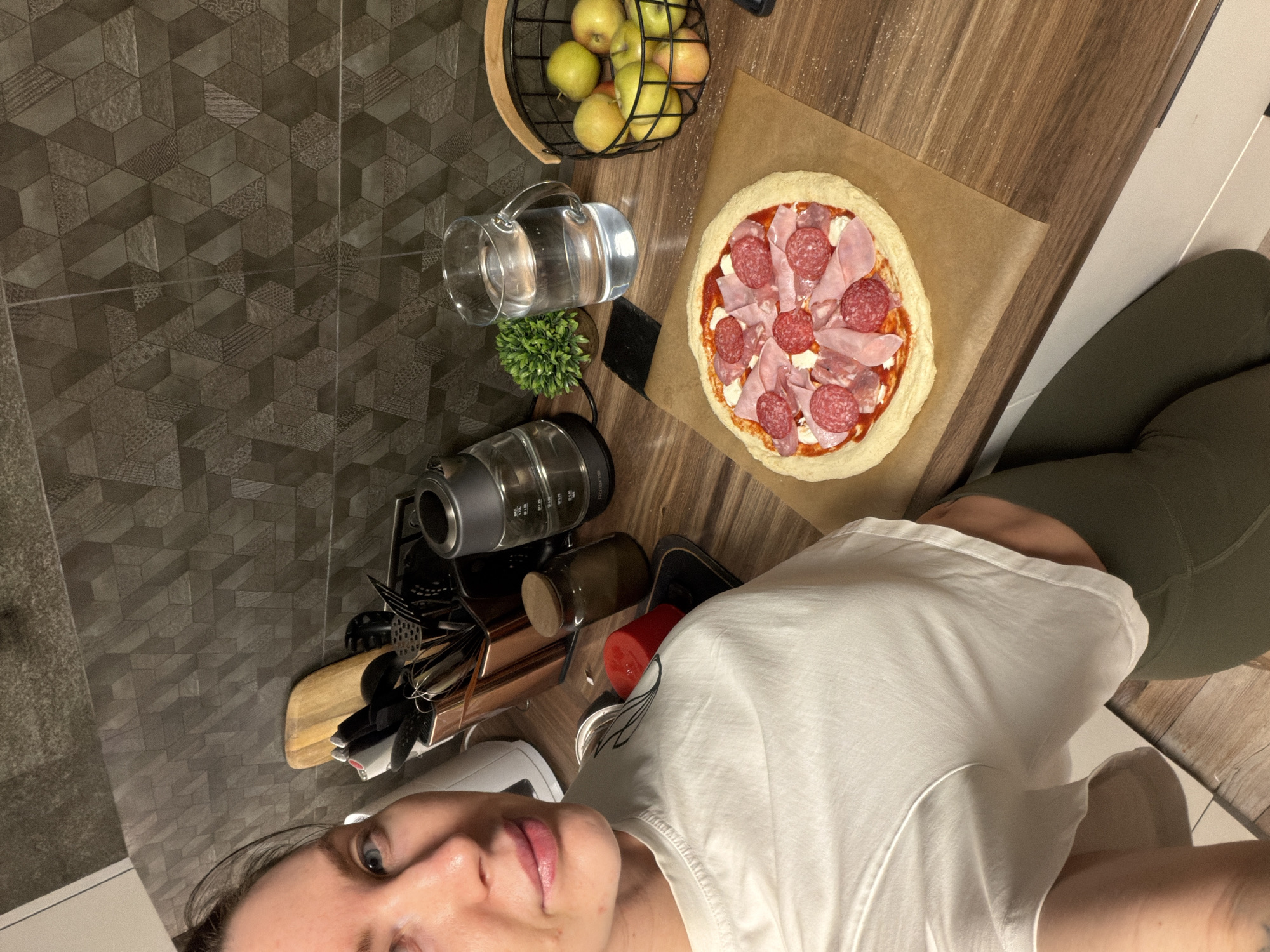A top-down selfie in what appears to be a kitchen in Perm, Russia, showing a person wearing a white shirt and olive-green leggings. Next to them is a pizza on parchment paper, topped with tomato sauce, cheese, salami, and ham. An apple basket with green and yellow apples, a pitcher of water, a small potted plant, and a utensil holder are also visible on the counter. The kitchen has dark grey patterned tiles on the wall and wood flooring. A kettle and other kitchen utensils are also on the counter.