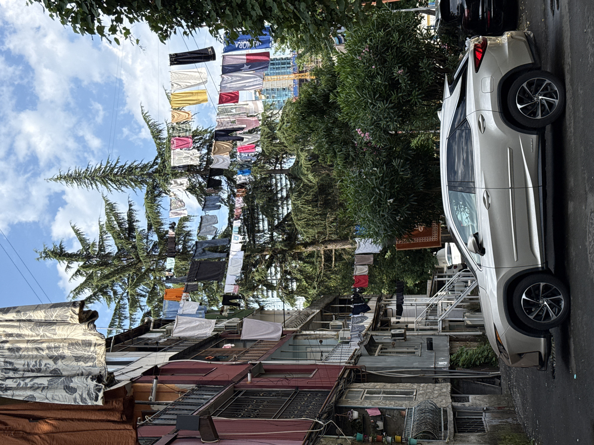 The image captures a street scene in an urban environment, likely in the city of Unknown, Unknown. In the foreground, a silver Lexus RX SUV is parked on a paved street. The vehicle is facing right, and its right side is prominently visible. Behind the car, a multi-story building with several balconies and windows stands to the left. Laundry is hung from clotheslines strung across the street and between buildings, with various garments drying in the open air. Tall evergreen trees are interspersed with the buildings, and some foliage is visible on the right side of the frame. In the background, a construction crane and a tall modern building can be seen, suggesting ongoing development in the area. The sky is partly cloudy. There are no people directly visible in the frame.