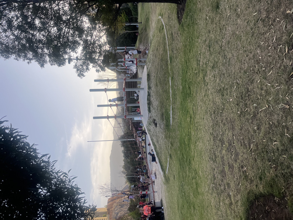 A park area with a playground and grassy field is visible. Children play on a tall, cylindrical playground structure connected with ropes. A mountain range is in the background, under a hazy sky. Trees frame the image on the sides. The field has a white painted semi-circular line on it.