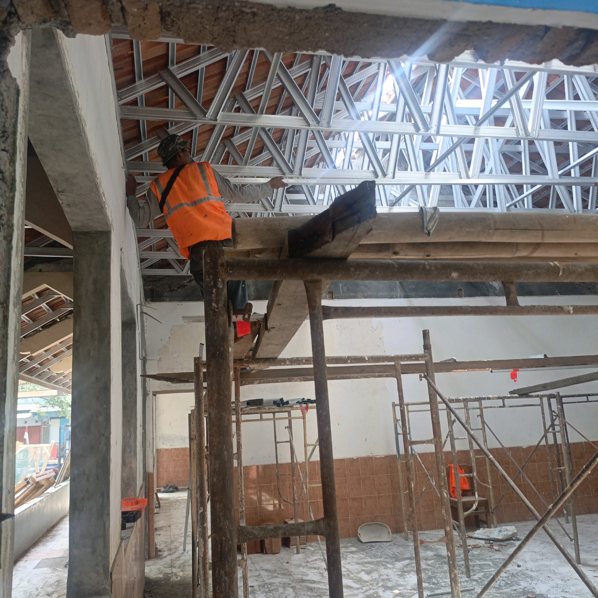 The image depicts an interior construction site, likely in Salatiga, Indonesia, with scaffolding and exposed roof trusses. A construction worker, wearing an orange safety vest and a camouflage hat, stands on scaffolding and appears to be working on the ceiling structure. The environment shows unfinished walls and floor, indicative of ongoing construction or renovation. Other visible objects include construction materials like wood planks, metal framing, and various tools. The lighting suggests it is daytime, with sunlight filtering through the roof openings. The overall scene conveys an active construction setting.