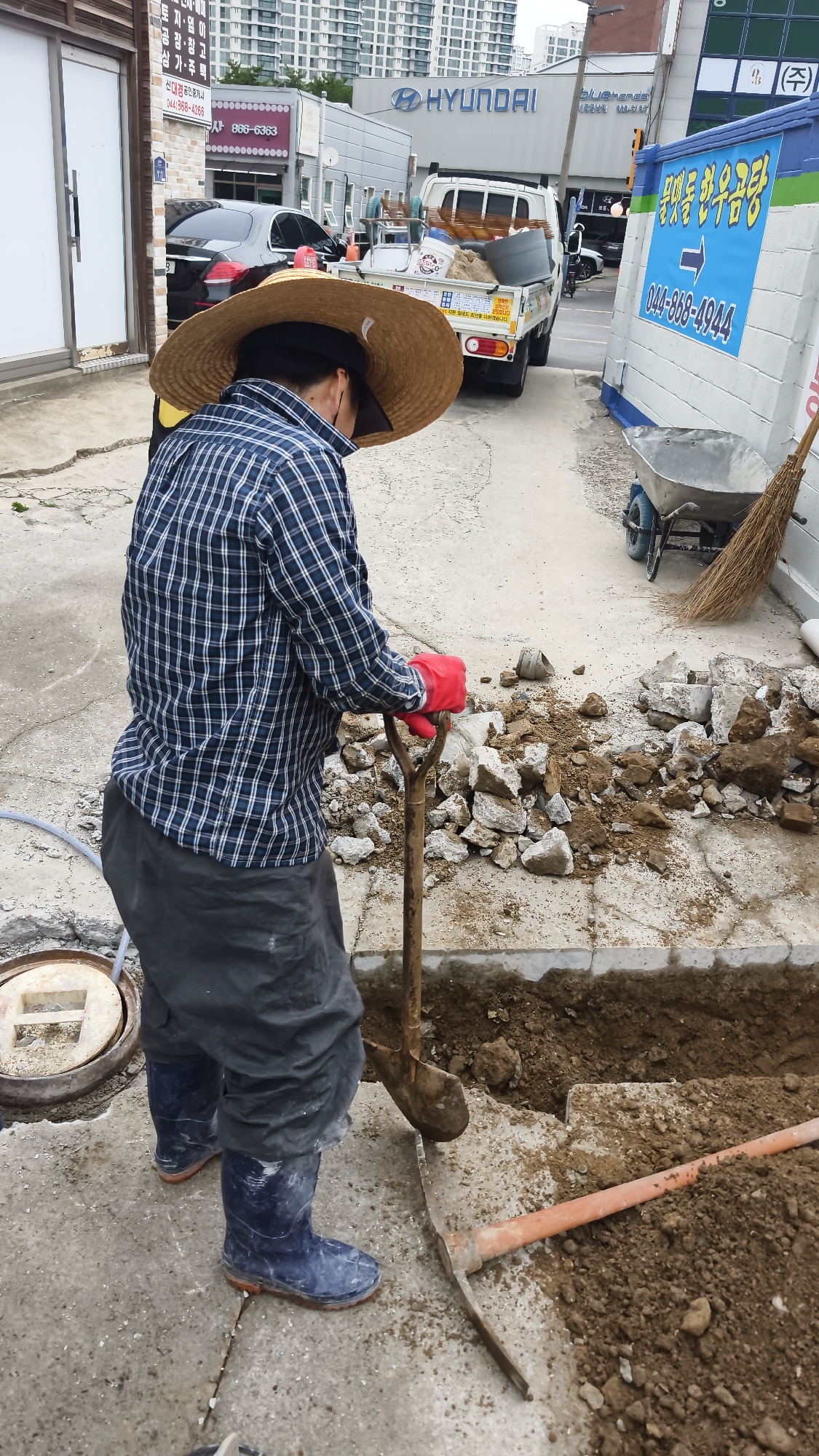 A person wearing a straw hat, a blue and white checkered shirt, grey pants, and blue rubber boots is using a shovel to dig a trench on a concrete surface. Red gloves are on their hands. The person is standing in what appears to be an outdoor work area in front of a building with "HYUNDAI" signage. There are piles of dirt and rocks near the trench, a wheelbarrow, a broom, and construction debris. A black car is parked in the background. The scene appears to be in the city of Sejong, South Korea, suggested by the Korean text on nearby signage.