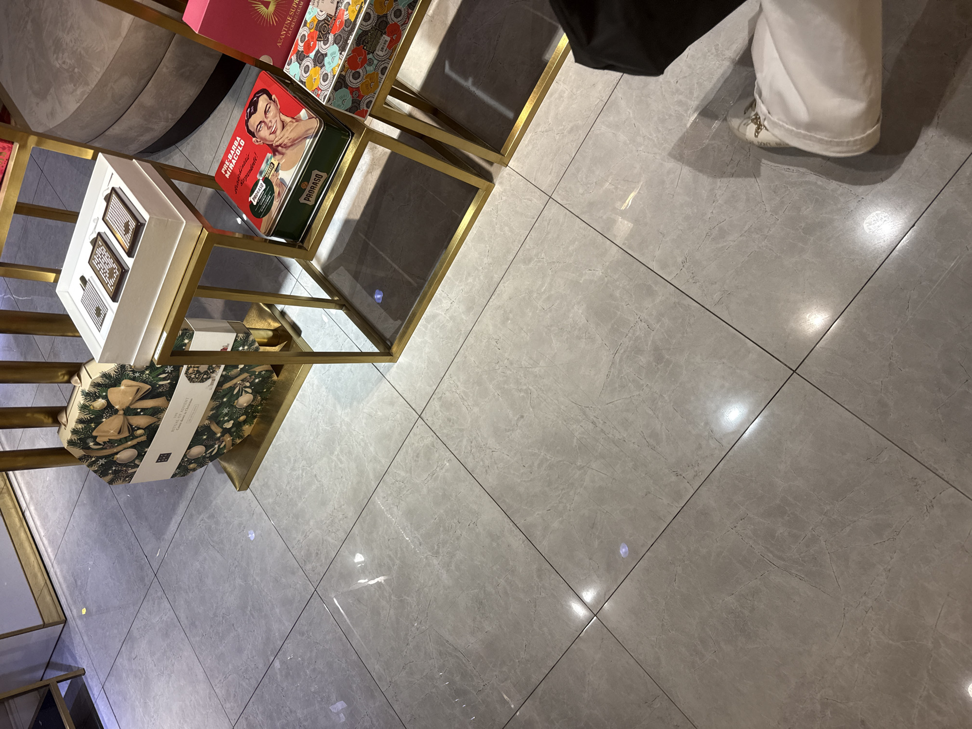 The image shows a retail display of cosmetic and grooming products, likely in a store. The items are arranged on gold-colored shelves. Prominently featured are several boxes and tins, including a green tin with a vintage-style image of a man and the text "PRORASO" and "PRE-BARBA MIRACOLO". Another item is a white box with smaller compartments containing what appear to be grooming products. A large, octagonal box adorned with a festive wreath and bow, labeled "RITUAL OF ADVENT", is also visible. In the background, out of focus, are other colorful boxes and the side of a large, upholstered circular structure.
The floor is tiled with large, light gray tiles that have a marble-like pattern, reflecting light from above. In the bottom right corner of the image, a person's lower legs and feet are visible. They are wearing white, loose-fitting pants and white slip-on shoes with gold embellishments. The overall impression is a well-lit retail environment, possibly a department store or a specialized beauty shop. The products suggest a focus on men's grooming and possibly holiday-themed gift sets. The lighting suggests it's daytime, with artificial lights adding to the illumination. The scene is clean and organized, typical of a retail display. There is no indication of the weather or a specific time of day beyond it being lit.