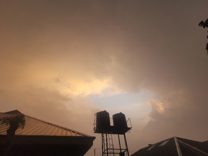 Under a turbulent, golden sky, two water tanks sit atop a metal frame, silhouetted against a gap of pale blue sky. Adjacent rooftops break the horizon line, also dark against the bright, overcast sky. A ladder runs up the side of the water tank structure, adding to the industrial feel of the scene, which is bathed in a warm, subdued light.