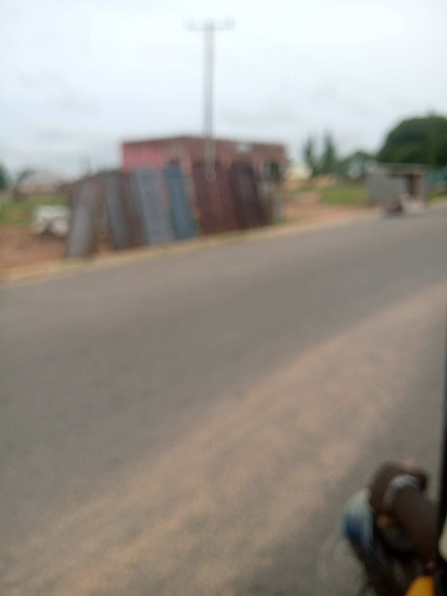 The image depicts a blurry outdoor scene, likely a street in Jalingo, Nigeria. In the background, a rectangular building with a red facade is visible, along with a tall, thin utility pole. To the left of the building, there appears to be a pile of metal sheets or corrugated iron, leaning against each other. A paved road occupies the foreground, with a portion of a person's head and shoulder, wearing a dark garment and a blue shirt, visible in the lower right corner, suggesting the photographer is in or on a vehicle. The sky is overcast.