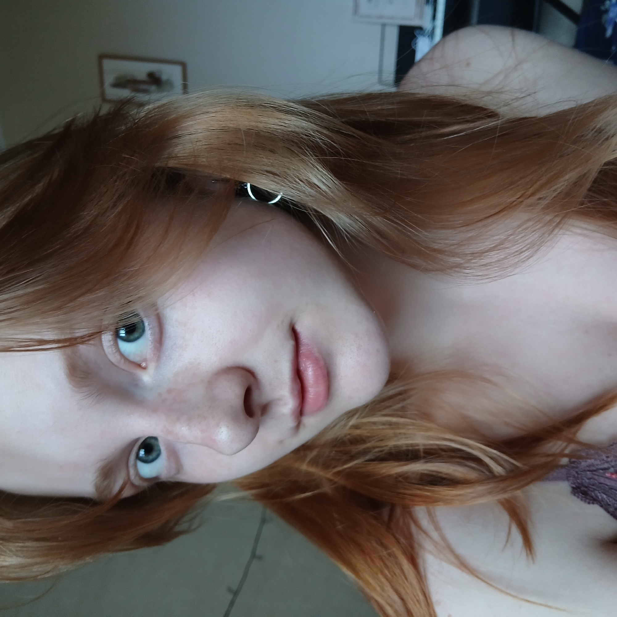 A close-up portrait, taken at a tilted angle, features a young person, likely female, with fair skin and numerous light freckles across her nose and cheeks. Her long, reddish-blonde hair cascades around her face and shoulders. She has striking blue-green eyes that look directly at the viewer, and her lips are a natural, soft pink.
A small, silver-colored hoop earring is visible in her left ear, partially concealed by her hair. The subject appears to be reclining or lying down, contributing to the casual and intimate composition of the photograph.
In the blurred background, a light-colored wall is visible, with a framed picture hanging indistinctly upon it. A glimpse of patterned, possibly knitted, purple fabric can be seen on the far right, suggesting clothing or bedding. The lighting is soft and diffused, creating an even illumination without harsh shadows, consistent with an indoor setting in Yekaterinburg, Russia. There is no visible text in the image. The overall impression is one of calm and natural beauty.