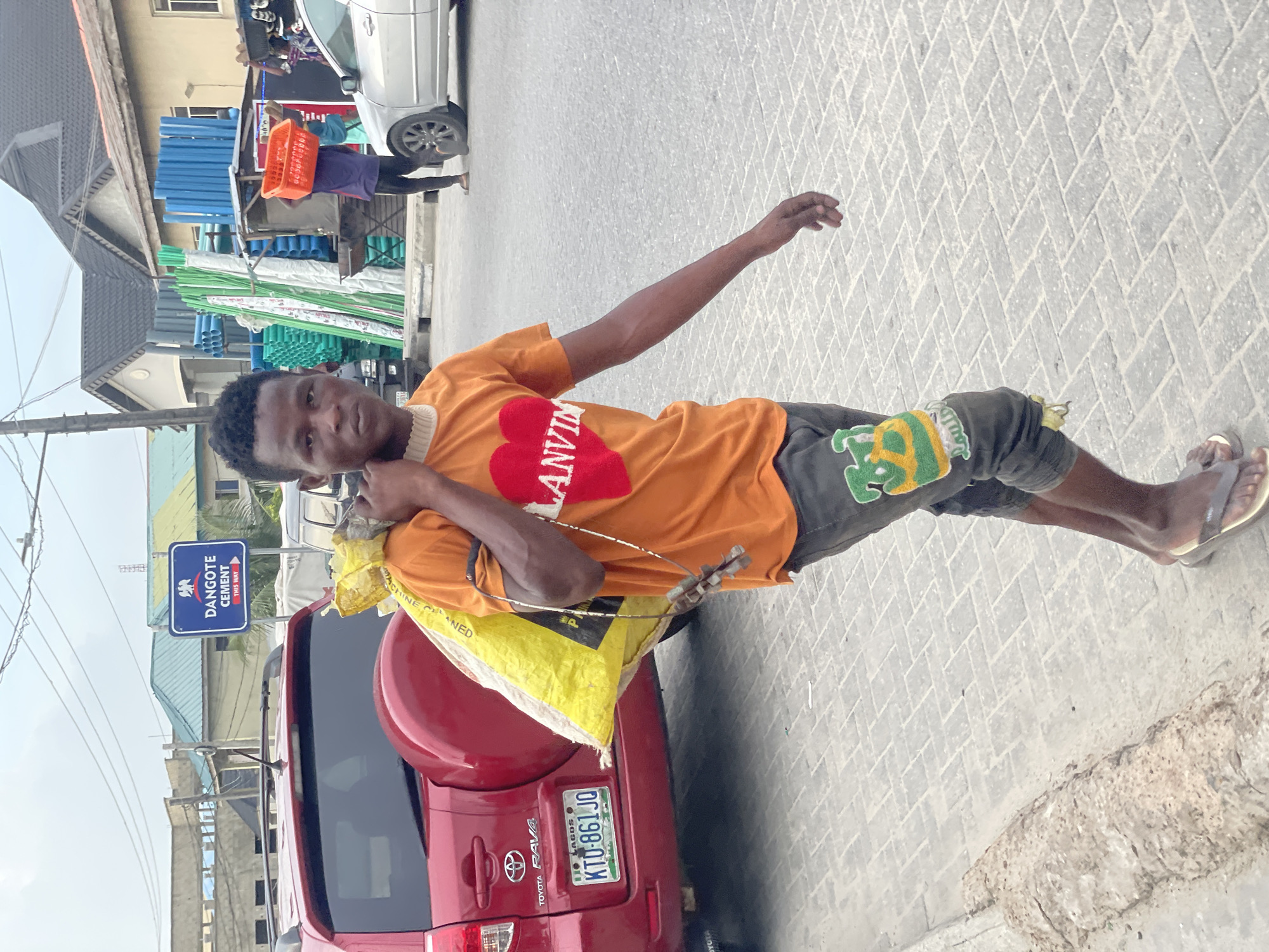 A young, dark-skinned man is the central subject in this daytime urban street scene in Ado, Nigeria. He wears an orange t-shirt with a red heart design bearing the text "LANVIN" and dark shorts with green and yellow motifs. He is carrying a yellow sack, partially labeled "MACHINE CLEANED," on his left shoulder and wears flip-flops. His right arm is outstretched, and he looks directly at the camera with a neutral expression.
Behind him is a red Toyota RAV4 car, displaying a Nigerian license plate "LAGOS KTU-861 JO". The ground is paved with light-colored interlocking blocks in a herringbone pattern. In the background, another person can be seen carrying an orange basket on their head, walking near a white car. Stacks of blue and green pipes and tubes are visible, indicative of a construction material supplier. A prominent blue rectangular sign points "THIS WAY" to "DANGOTE CEMENT". Buildings with grey roofs line the street under bright, clear skies.