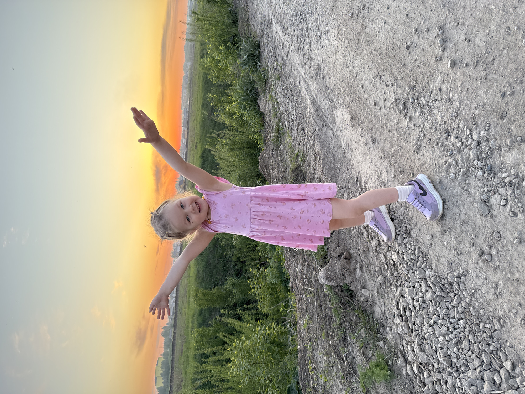A young girl stands with her arms outstretched against the backdrop of a sunset in Горный, Russia. She wears a light pink dress with a floral pattern and light purple sneakers with white socks. The ground is a mix of dirt and small rocks, and there are green bushes in the background. The sky transitions from yellow to orange near the horizon, suggesting either sunrise or sunset.