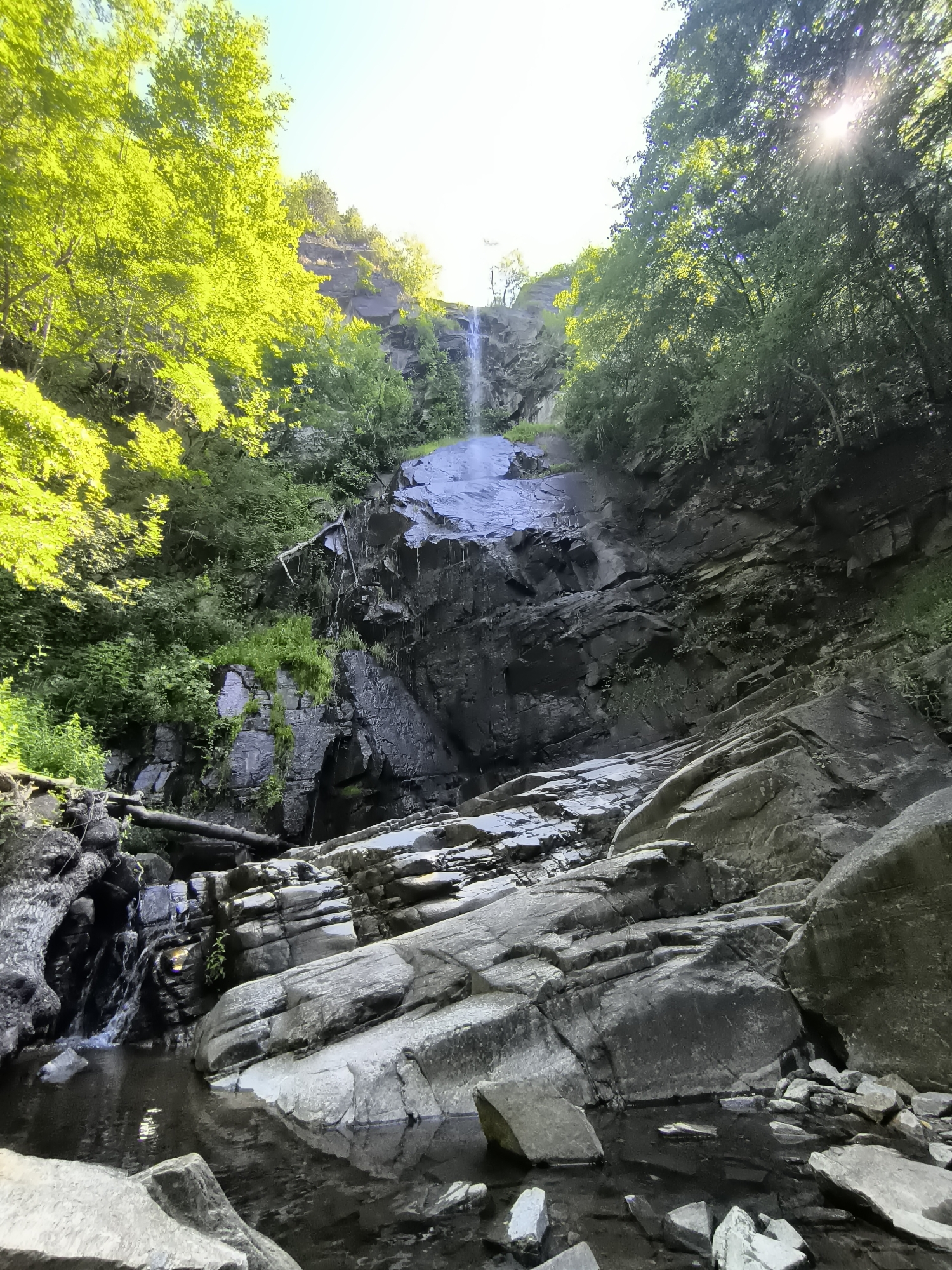 This is a vertical, eye-level, full shot of a cascading waterfall in a rocky, wooded environment. The waterfall is the focal point, with water flowing down a dark, wet rock face. The surrounding area is densely covered with lush green trees and foliage, with the bright sun shining through the canopy, casting dappled light. Large, gray rocks are prominent in the foreground, some submerged in the dark water at the base of the waterfall. A fallen log lies across some of the rocks near the water. The scene evokes a sense of natural beauty and tranquility.