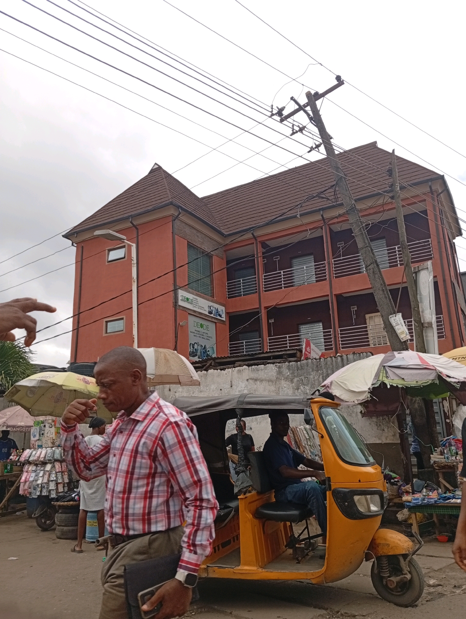 This is a medium-shot, eye-level, slightly tilted outdoor photograph taken in Akowonjo, Nigeria. The focal point is a three-story, salmon-colored building with a brown tiled roof, featuring multiple balconies with white railings. Power lines crisscross the sky above the building. In the foreground, a man wearing a red and white checkered shirt and khaki pants walks towards the right, holding a black folder and a smartphone. He is wearing a wristwatch. To the left of the man, a yellow auto-rickshaw is parked, with a man in a blue shirt and jeans driving it. Two street vendors with umbrellas are visible behind the auto-rickshaw. A hand is pointing from the left edge of the frame. The ground appears to be a dirt or paved road. The sky is overcast.