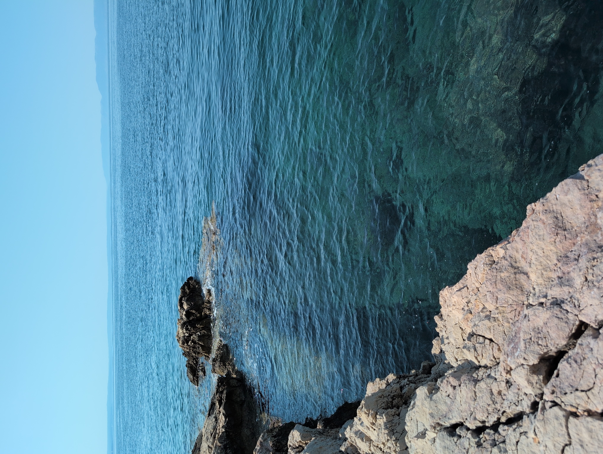 The image depicts a tranquil coastal scene from Kostrena, Croatia. In the foreground, a rugged, light-colored rocky shoreline dominates the left side, with textured, cracked surfaces. Beyond the rocks, the crystal-clear, turquoise water of the Adriatic Sea stretches out towards the horizon. The water is gently rippled, and sunlight penetrates to reveal the submerged rocks and seabed beneath. In the distance, a faint, hazy outline of a coastline or islands can be seen against the pale blue sky, suggesting a clear and calm day. There are no people or animals visible in the frame, emphasizing the serene and untouched nature of the environment. The overall impression is one of natural beauty and peacefulness, characteristic of the Croatian coastline.