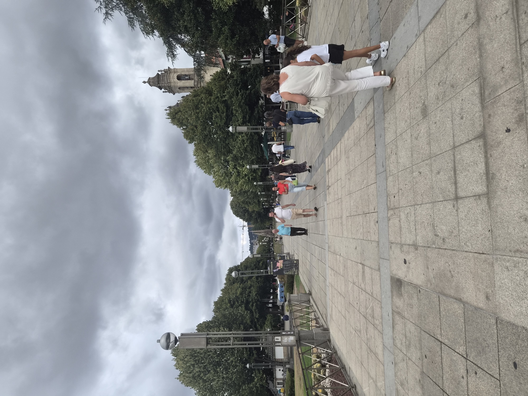 A wide, paved walkway lined with trees and lampposts is the setting for this image. Several people are walking along the path. In the background, a bell tower of a church or cathedral rises above the trees, characteristic of the cityscape of Braga, Portugal. The sky is overcast, suggesting a cloudy day. The walkway is constructed of large, square stone tiles, with a narrower stone strip running down the center. A metal railing with a decorative pattern separates the walkway from a grassy area on the left. Several lamp posts with spherical tops are visible along the walkway.