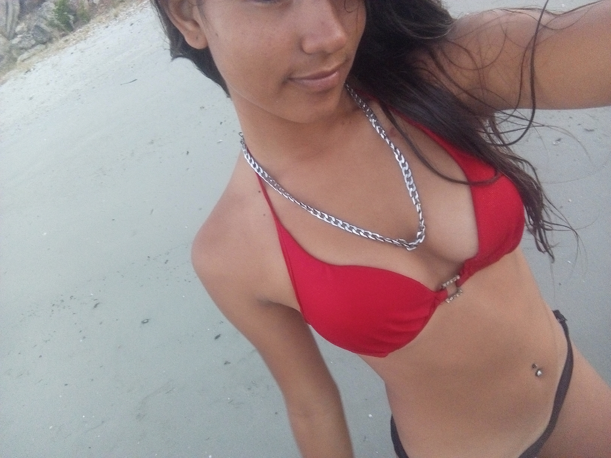 The image is a selfie of a young woman at a beach. She has dark hair and is wearing a red bikini and a silver chain necklace. She also has a navel piercing. The beach appears to be sandy and the lighting suggests it is daytime. The location is the city of Juan Griego, Venezuela.