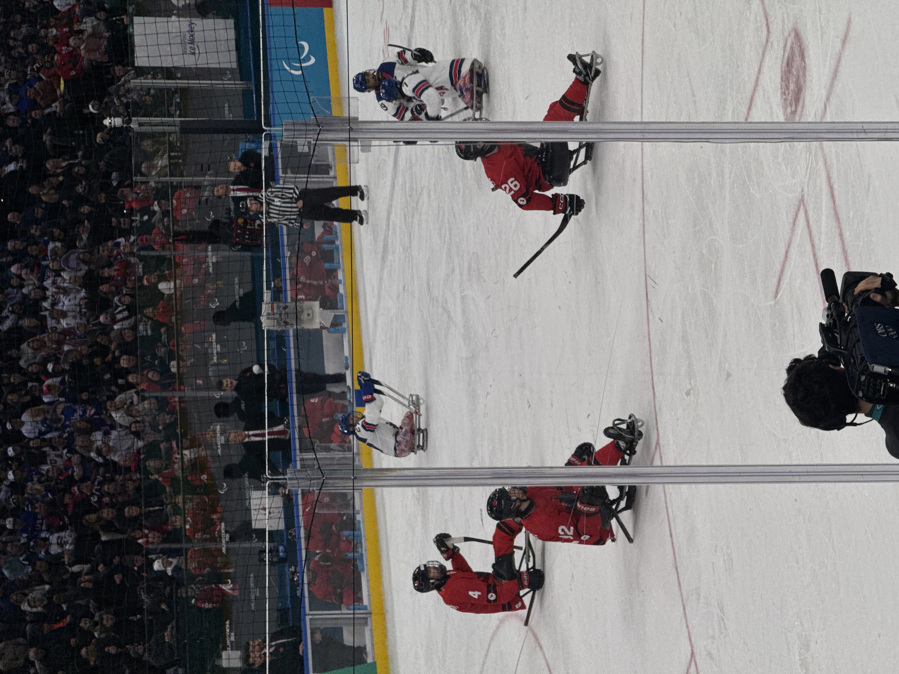 This image captures a sled ice hockey event inside an indoor ice arena, identified by prominent "MILANO CORTINA 2026" and Paralympic symbol signage, indicating its association with the 2026 Winter Paralympic Games in Milan, Italy. The central and lower frame features the white ice rink surface with faint red markings. Six athletes are actively positioned on the ice, each seated on a specialized sled with attached skates, wearing helmets, protective gear, and using two short hockey sticks for propulsion and puck control. Four players are in red uniforms, with visible numbers '4', '12', and '26' on their backs. Two players are in white, blue, and red uniforms. One referee in a black and white striped shirt stands on skates near the left boards. In the immediate foreground, a camera operator, viewed from behind, is operating a professional video camera with "OBS" visible on its cover. The background consists of dark, tiered spectator stands, filled with an audience, partially obscured by black protective netting. Blue and dark grey rink boards display "MILANO CORTINA 2026", the white Paralympic "agitos" symbol, and a "World Para Ice Hockey" sign.