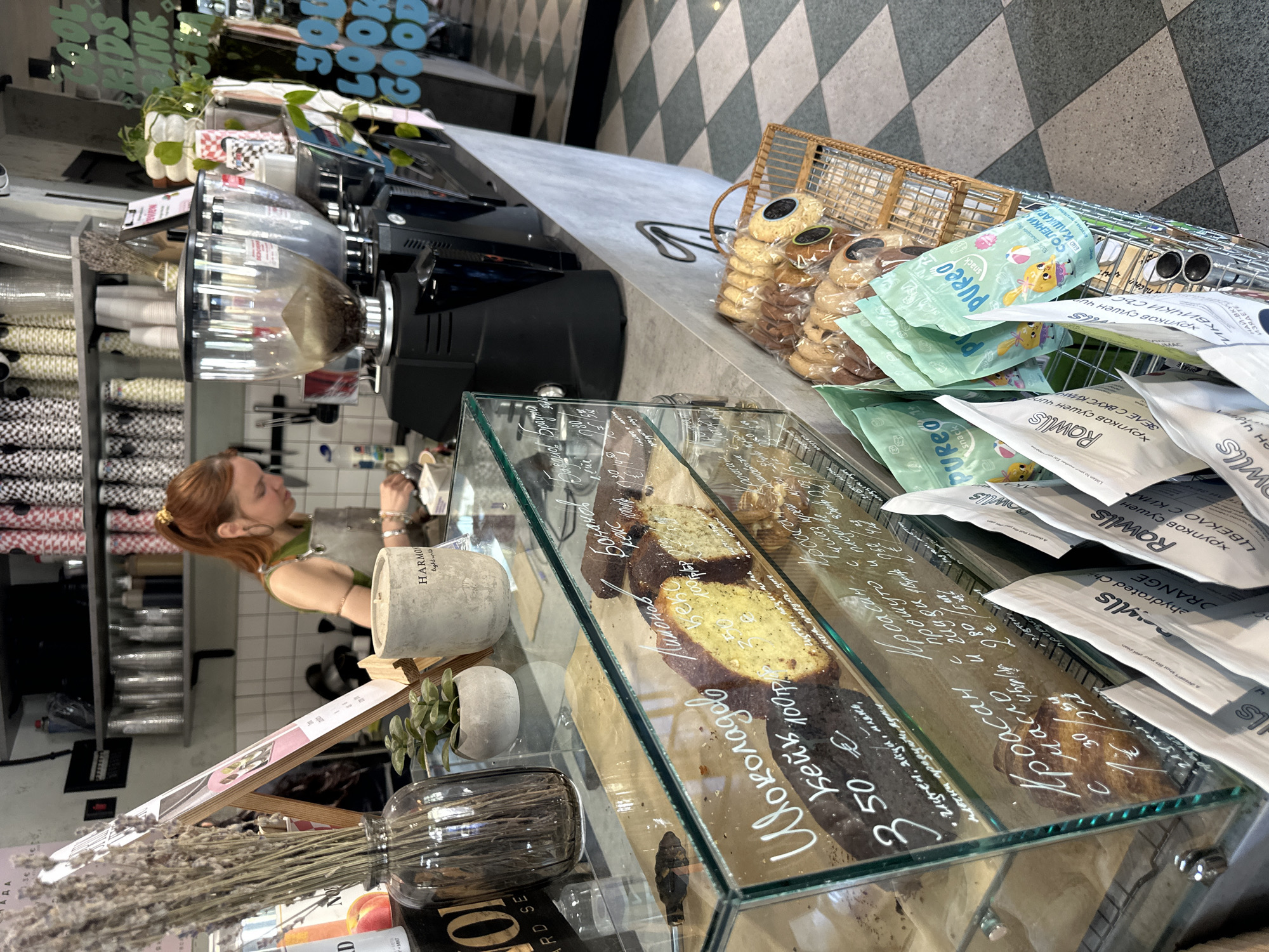 The image depicts the interior of a café or bakery in Sofia, Bulgaria. A woman with reddish-brown hair is visible behind the counter, appearing to be preparing or serving items. In the foreground, there's a display case filled with pastries, including chocolate cake and croissants, with handwritten pricing and descriptions in what appears to be Bulgarian or a similar Cyrillic script. To the right of the pastries, there's a basket holding packages of cookies and bagged snacks from brands like "PUREO" and "Rowlls," also with Cyrillic text on the packaging. The floor is a checkered pattern. A coffee grinder and other equipment are visible on the counter behind the display case. Decorations include plants and dried flowers. The environment suggests a modern, stylish café with a focus on fresh baked goods and snacks.