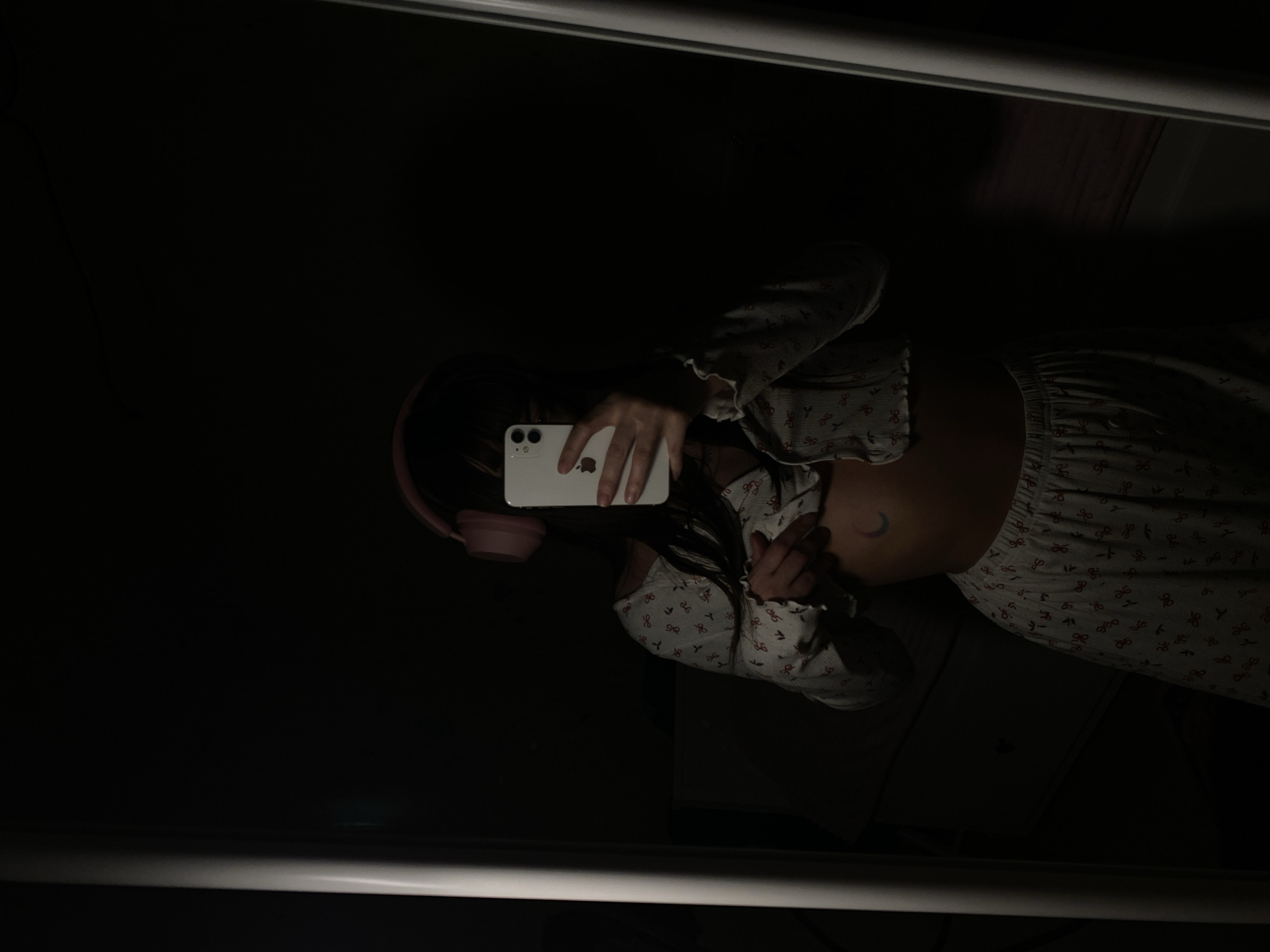 A mirror selfie shows a young woman with long dark hair, wearing pink headphones. She is holding a white smartphone to take the picture. She has on a matching pajama set patterned with small red bows. A small crescent moon tattoo is visible on her stomach. The background appears dark and obscured. There is no evidence to suggest the photo was taken in Votkinsk, Russia.