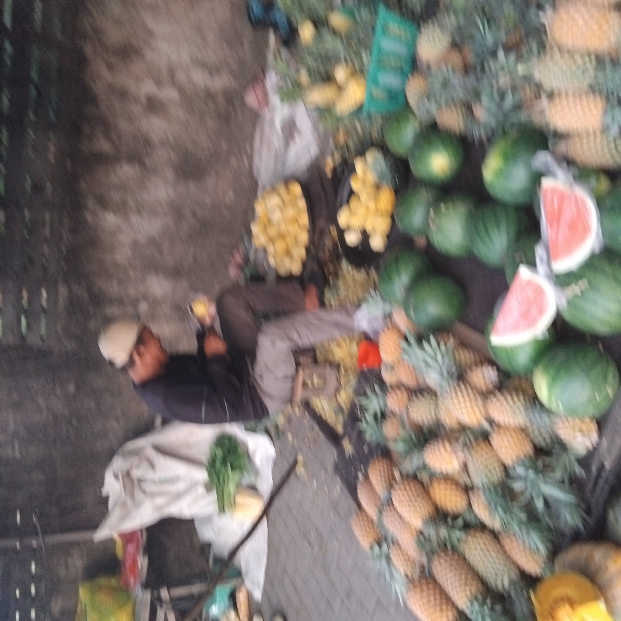 A man wearing a light-colored cap and a dark long-sleeved shirt, along with brown pants, is seated on a low wooden stool and peeling a pineapple. He is surrounded by a variety of fruits displayed on the ground. In front of him, to his left, are piles of pineapples. To his right, there are two large, round, shallow containers overflowing with yellow fruit, possibly also pineapples or another type of fruit. Further to the right, and in the foreground, are several green watermelons, some of which have slices of watermelon on display. In the background, a white sheet or tarp is spread out, with some green leafy vegetables placed on it. The scene appears to be an outdoor market stall. The overall setting is somewhat blurry, suggesting movement or low light conditions. This scene is located in a city in Banjarbaru, Indonesia.