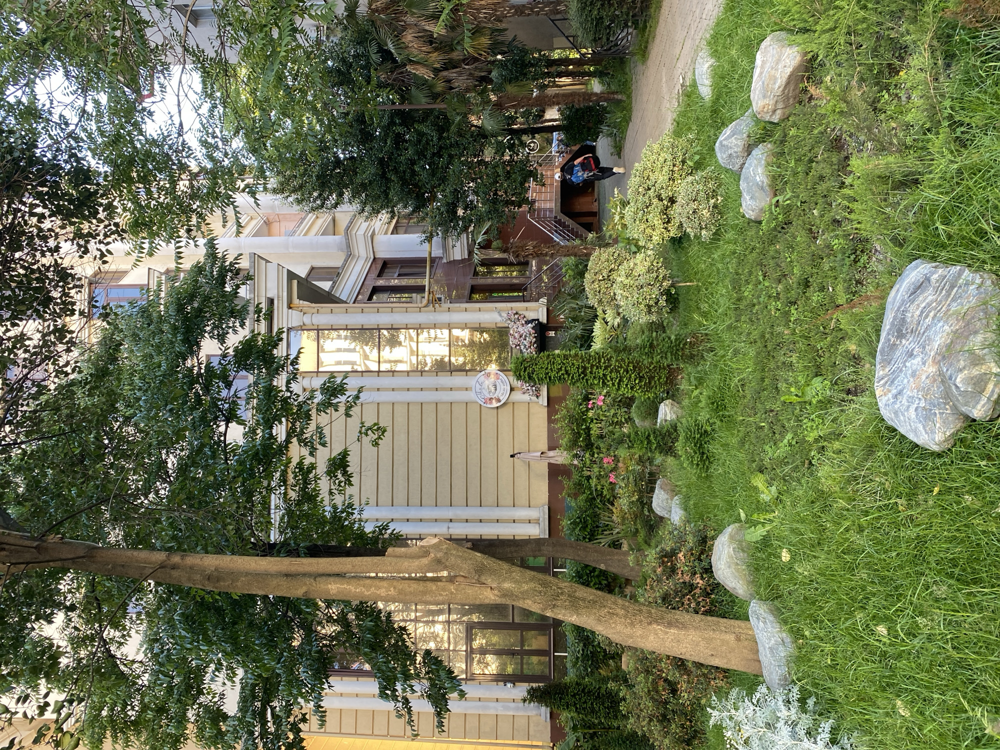 The image captures a vibrant outdoor scene featuring a multi-story building alongside a lush garden, likely in Sochi, Russia, given the context. The building has a cream-colored facade with intricate architectural details, including decorative molding and large windows that reflect the sky and surrounding greenery. A prominent set of stairs with metal railings leads to a lower entrance of the building, where a person is visible. This individual is wearing a dark jacket and a blue backpack, appearing to be crouched or seated near the bottom of the staircase.
The foreground and mid-ground are dominated by a meticulously maintained garden, characterized by dense green grass, various shrubs, and several large, smooth gray rocks. A variety of trees, including tall palm trees and other deciduous types with abundant foliage, frame the scene, providing shade and depth. Sunlight brightens the scene, creating reflections on the building's windows.
Affixed to the building's facade, above an outdoor umbrella and amidst flowering plants, is a round sign with a light background featuring what appears to be a stylized image or logo, though specific text is not clearly legible. The overall impression is one of a well-kept, serene environment, possibly a residential area or a hotel entrance. The time of day appears to be daytime, with clear weather.