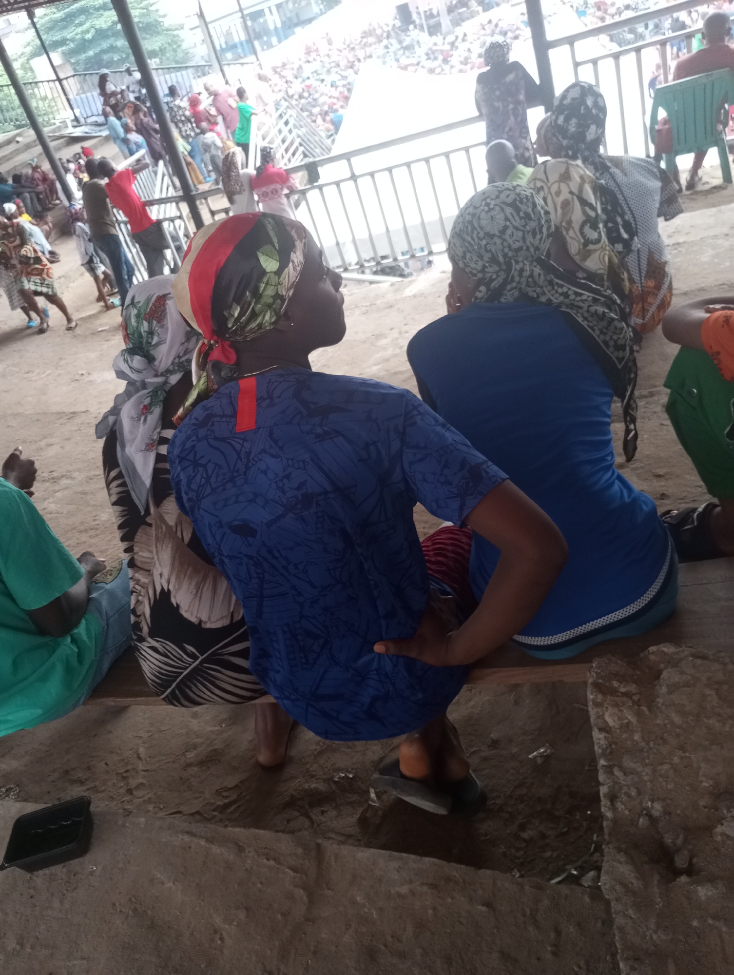 In a gritty outdoor urban setting, potentially a marketplace or gathering area in Unknown, Unknown, several individuals are captured in a moment of repose. In the foreground, a person wearing a vibrant blue garment with a patterned design sits on a wooden bench, their left hand resting on their thigh. Adjacent to them, another individual in a white and black patterned dress is also seated. To the left, a person in a bright green shirt is visible.
In the middle ground, a crowd of people is gathered behind a metal railing, suggesting an event or a public space. Many are dressed in colorful attire, with some wearing headscarves. The background reveals a densely populated area with buildings and structures, hinting at a bustling urban environment. The ground is a mixture of dirt and possibly concrete. A black plastic container is present on the ground in the lower left quadrant of the image. The lighting suggests an overcast day.