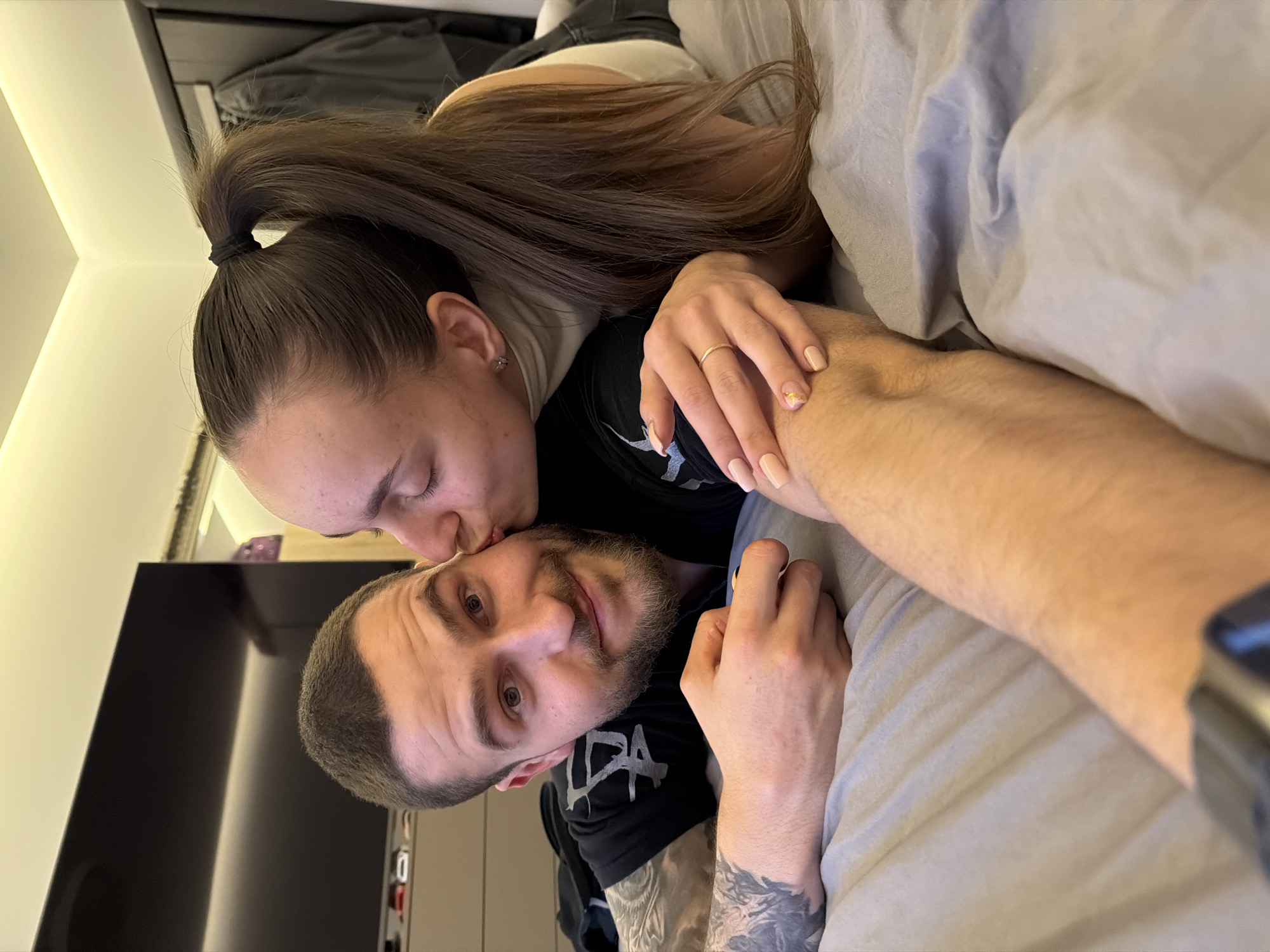 A couple is in bed. The woman, with her hair in a ponytail, is kissing the man on the forehead. The man has a beard and tattoos on his arm. They are lying on grey bedsheets. A television is visible in the background, along with a closet door. The image provides insufficient information to determine the location as Sofia, Bulgaria.