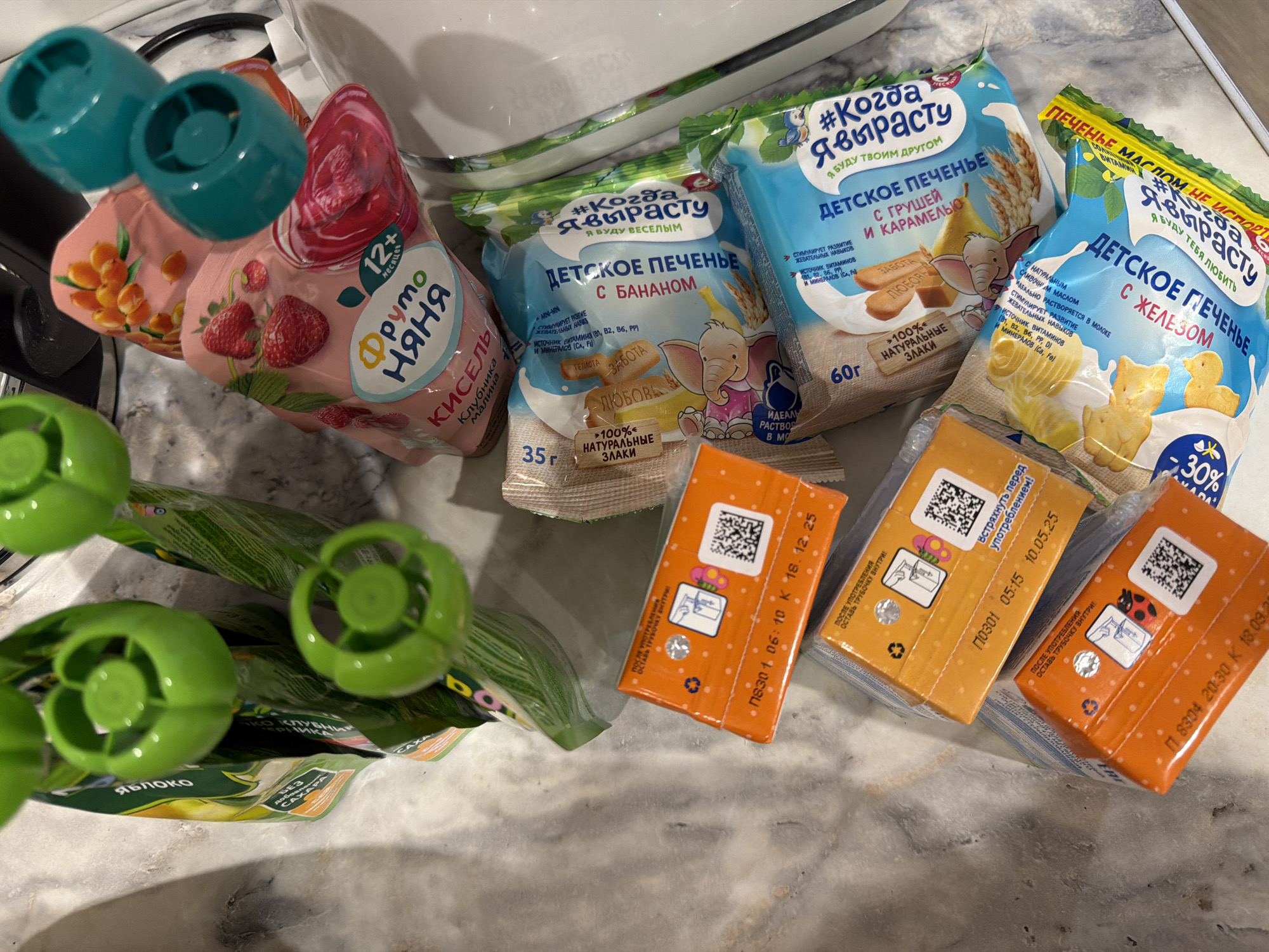 The image shows an assortment of baby food items on a gray countertop, likely in a kitchen. The items include: two pouches of "Fruto Nyanya" kissel drink, one is strawberry-raspberry flavor, the other is sea buckthorn flavor; three packages of "Kogda Ya Vyrastu" baby cookies in banana, pear-caramel and iron-enriched milk flavors; two green pouches of apple puree; and three small orange juice boxes. The juice boxes have expiration dates printed on them. The image appears to be taken from a top-down perspective. The objects are likely arranged for consumption or a product display. There are no people visible. There is no indication that this image was taken in Rostov-on-Don, Russia.