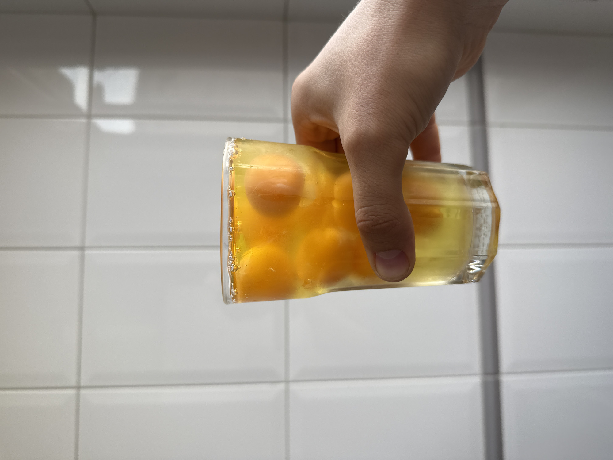 A hand holds a clear glass filled with egg yolks and whites against a background of white tiled walls. The scene lacks specific details that would place it in Gijón/Xixón, Spain.