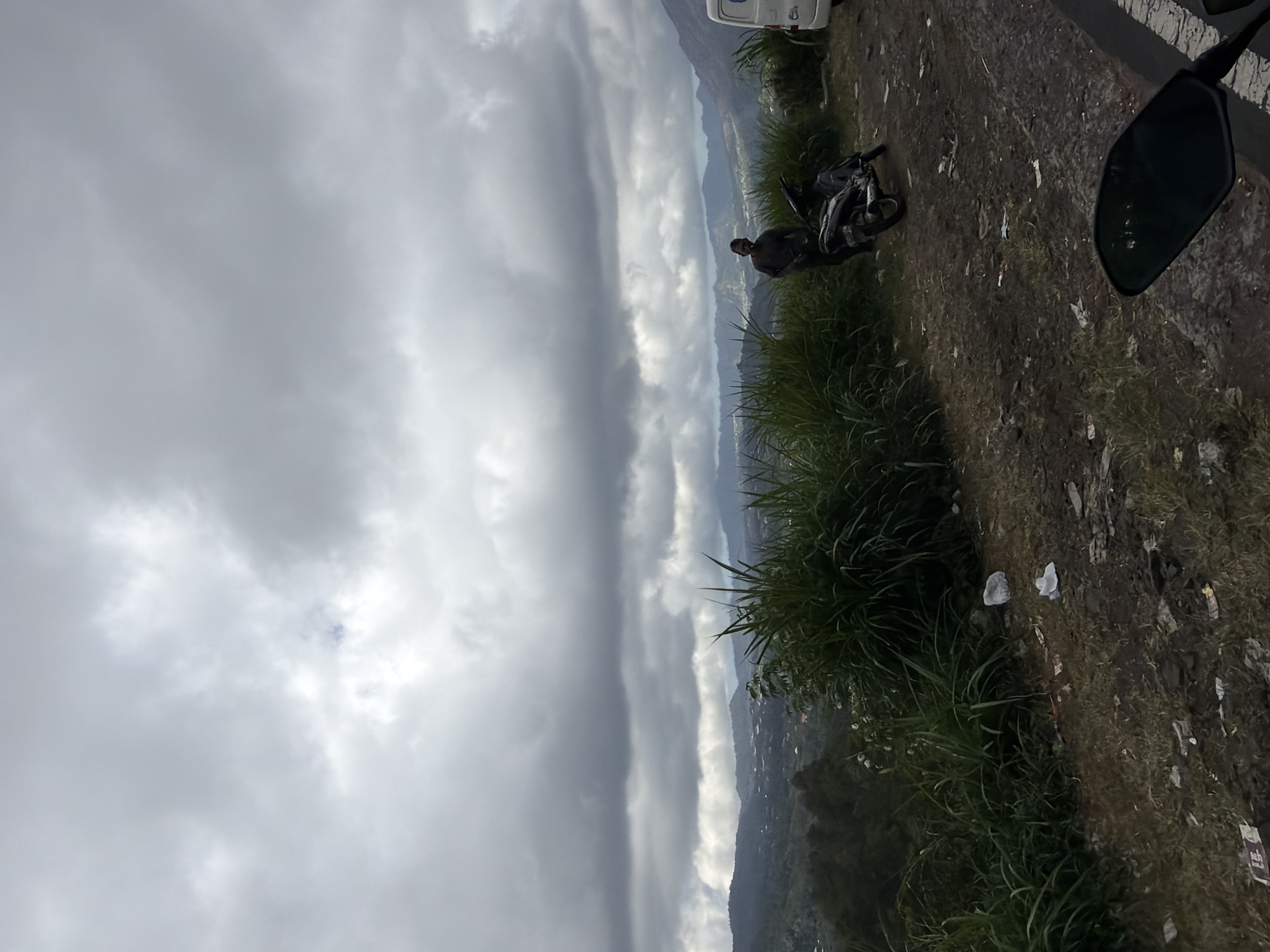 The image depicts a scenic overlook in Kintamani, Indonesia, characterized by a dramatic, cloudy sky and a lush green landscape. In the foreground, a person stands next to a parked motorcycle. The ground is rough and unpaved, with some scattered litter. The background reveals rolling hills and distant mountains, suggesting a rural or mountainous setting. The overall atmosphere is somewhat somber due to the overcast weather.
