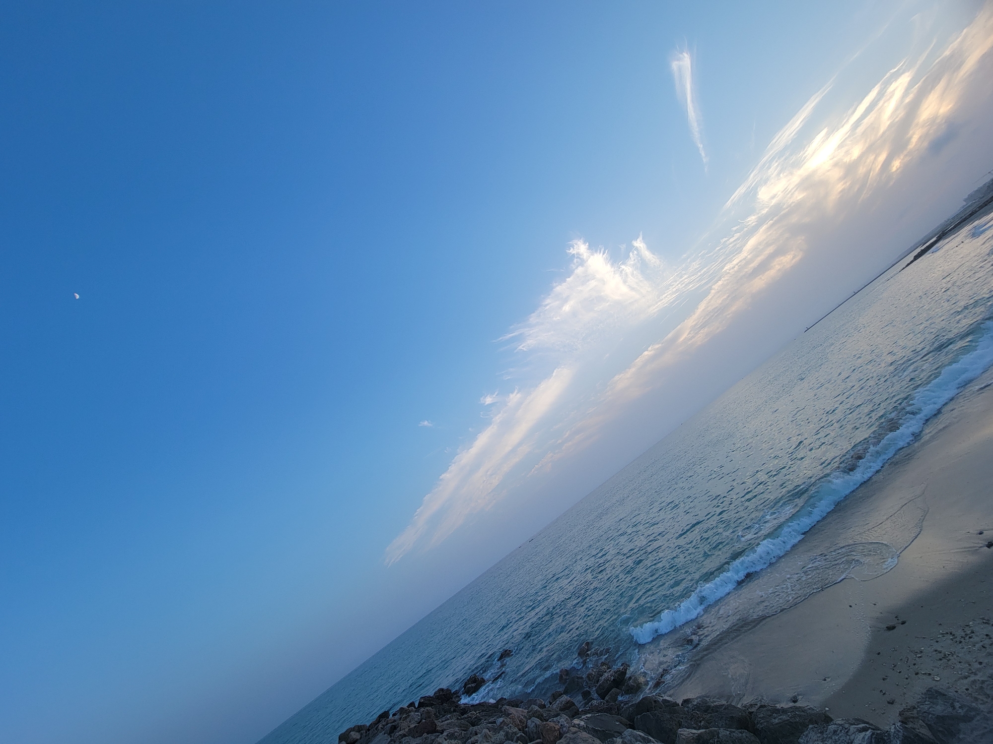 The image depicts a tranquil beach scene at Quarteira, Portugal, under a clear blue sky. A sliver of the moon is visible in the upper left quadrant. Wispy white clouds streak across the sky, illuminated by the soft glow of sunlight filtering from the right side of the frame. The turquoise ocean gently laps the sandy shore, with small waves breaking near the waterline. The foreground is composed of dark, rough-hewn rocks, suggesting a breakwater or natural rocky outcrop. The overall impression is one of peaceful solitude, bathed in the warm light of early morning or late afternoon. No people are visible in the scene.