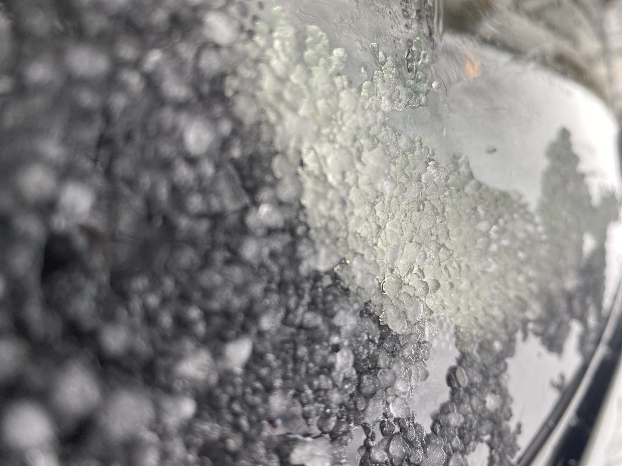 The image shows a close-up view of ice formations on a glass surface, likely a car window. The ice appears to be granular and partially melted, obscuring the view beyond. Through the clearer patches of glass, a blurry image of trees and other objects can be seen, suggesting an outdoor environment. There is no indication of the city of Chaikovsky, Russia or any people, actions, or interactions visible within the frame.