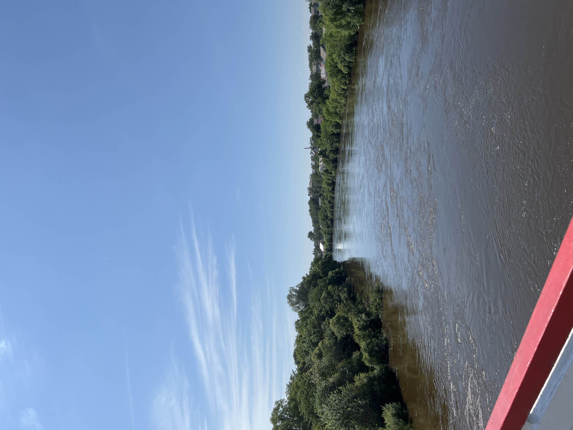 The image shows a wide, muddy river flowing through a sparsely populated area under a clear blue sky with wispy white clouds. Lush green trees line both banks of the river, with a few houses and buildings visible in the distance on the right bank, suggesting a small town or village. The foreground shows a red and white railing, possibly from a bridge or overlook, from which the photograph was taken. The overall impression is a tranquil, natural landscape with a hint of human settlement. The location is identified as Khor, Russia.