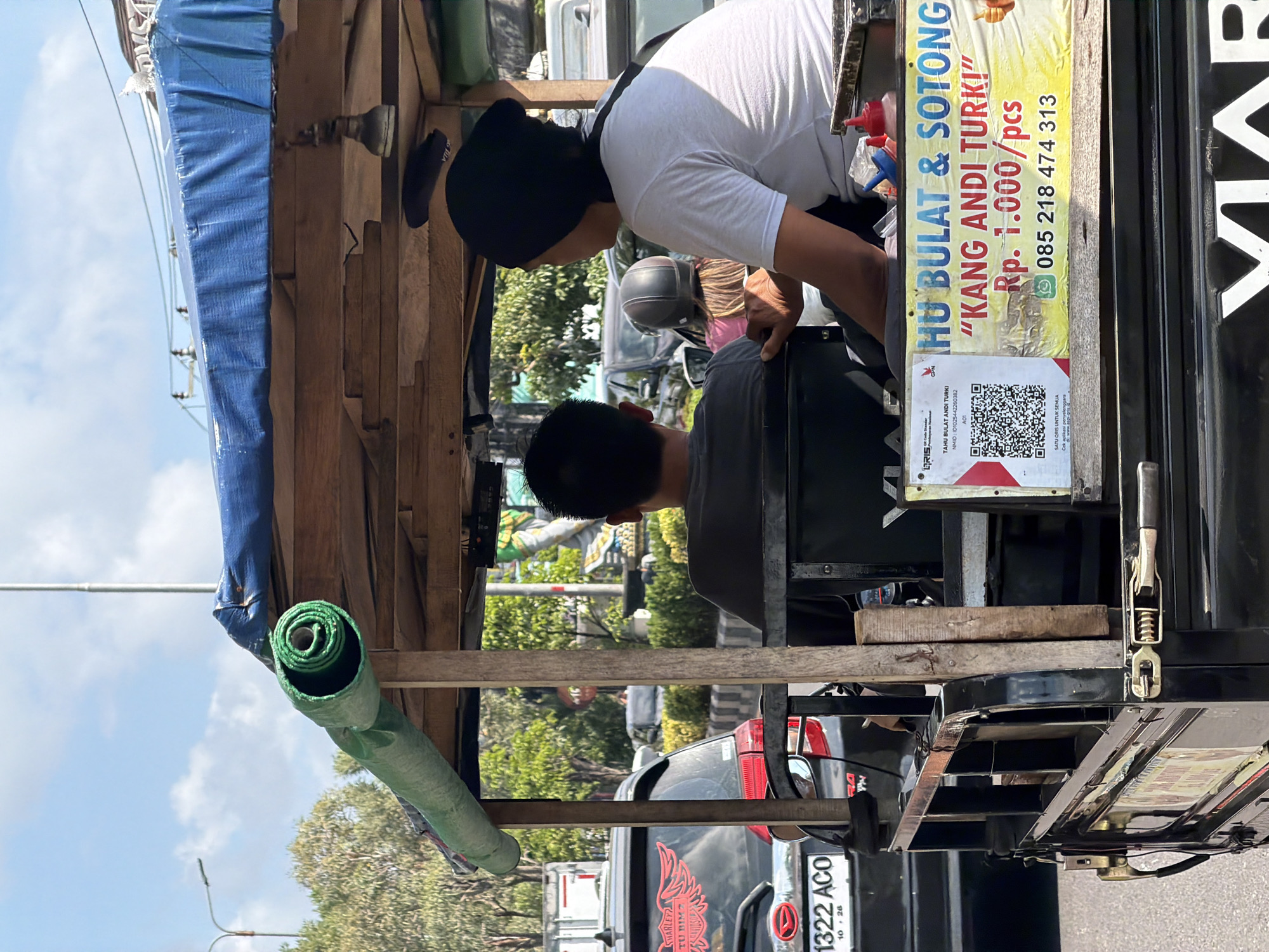 In Kuta, Indonesia, a modified open-air vehicle, possibly a type of auto-rickshaw or small truck, is pictured on a street during the daytime. The vehicle is partially covered by a blue tarp and a rolled-up green tarp. Inside, two individuals are visible. One, wearing a black beanie and a white t-shirt, is seated and facing away from the camera. Next to them, another person in a dark shirt is also seated, with their back to the camera. The vehicle displays several signs, including one with a QR code and text in Indonesian, which appears to be advertising a food vendor or service. The background reveals a street scene with other vehicles, trees, and buildings under a bright, partly cloudy sky. A black car with a red decal on its rear is parked nearby. The overall impression is of a bustling urban environment.