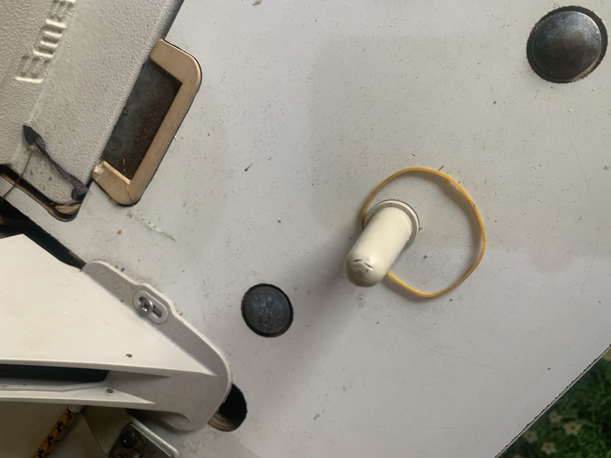 The image shows a close-up, overhead view of a sewing machine's surface. A cream-colored plastic component, possibly a part of the machine's housing or a lever, is visible on the left. A single yellow rubber band is looped around a white cylindrical object, also likely part of the sewing machine, positioned in the lower-middle section of the frame. The surface of the sewing machine is a textured, light gray material. Several screws and indentations are present on the surface. The background is out of focus, but hints of green and floral patterns suggest a domestic setting. The image is devoid of any human presence or activity. The location of Jauro Yinu, Nigeria is not depicted in this image.