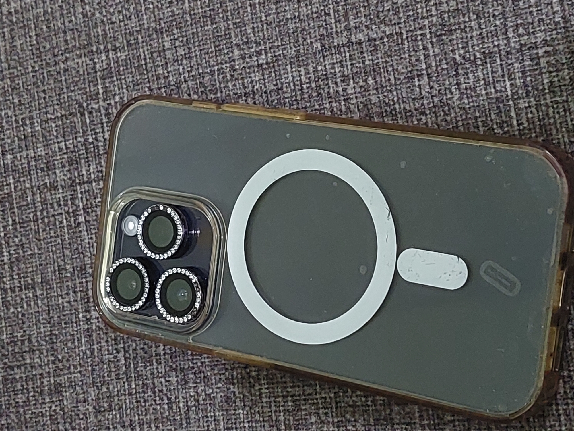 The image shows the back of a dark-colored smartphone with a clear case that has a subtle brown tint on the edges. The phone has a triple-lens camera system on the upper left corner, each lens surrounded by a ring of small, clear gemstones. A white circular magnetic charging ring is visible in the center of the phone's back, with a smaller white oval-shaped component situated below it. The phone is placed on a dark, textured surface that appears to be fabric, possibly a tablecloth or a carpet. There are no people or any other discernible objects or activities in the image. The location is not specified or implied in the image, and therefore cannot be determined as being in the city of Dumai City, Indonesia.